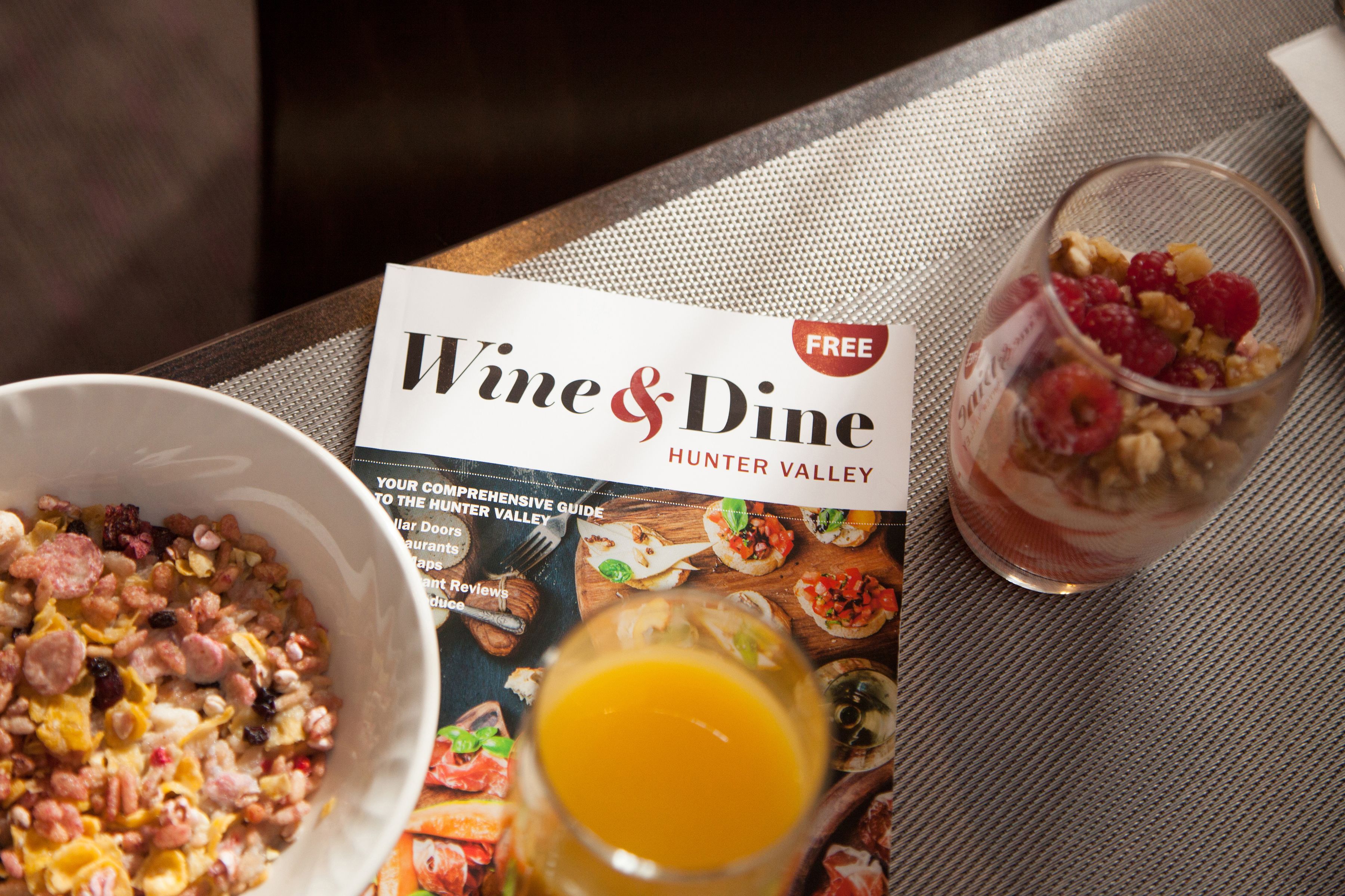Une table avec une brochure intitulée « Vins et gastronomie de la Hunter Valley », un bol de céréales avec des noix et des raisins secs, et un verre de jus d’orange.