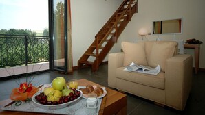 Un salon avec un escalier en bois, un canapé, une table à manger ornée de fruits et de pâtisseries, et une vue sur la verdure à travers une porte vitrée.
