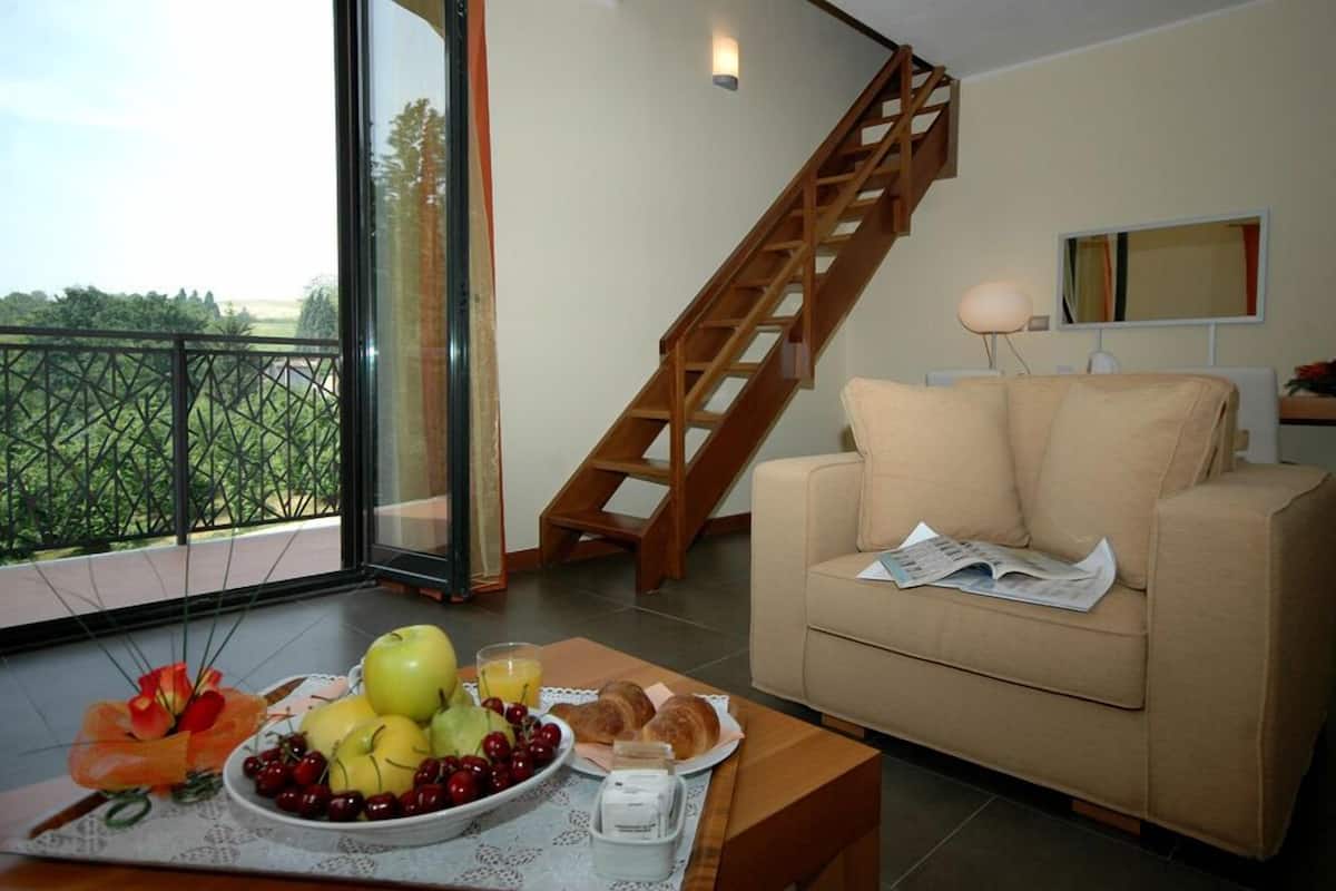 Sala de estar com escada de madeira, um sofá, mesa de jantar com frutas e doces, e vista para o verde através de uma porta de vidro.