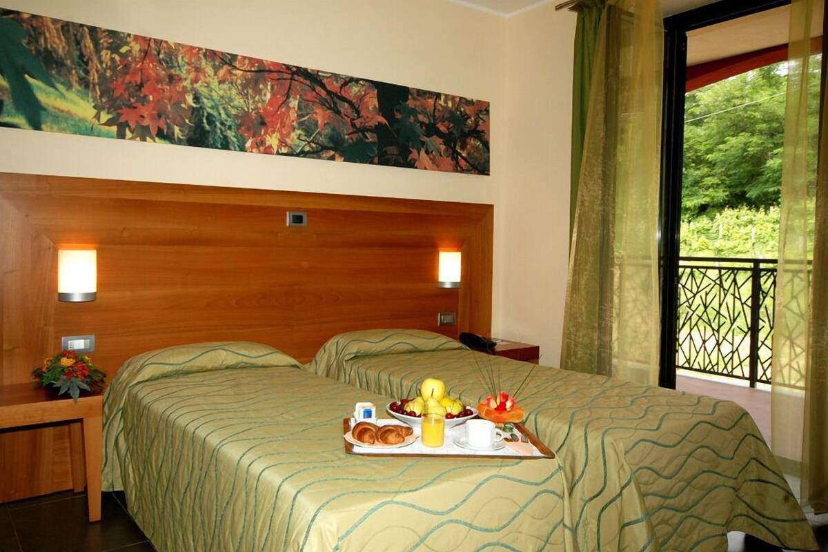 Quarto de hotel com duas camas, cabeceira de madeira, pintura acima das camas e varanda com vista para o verde.