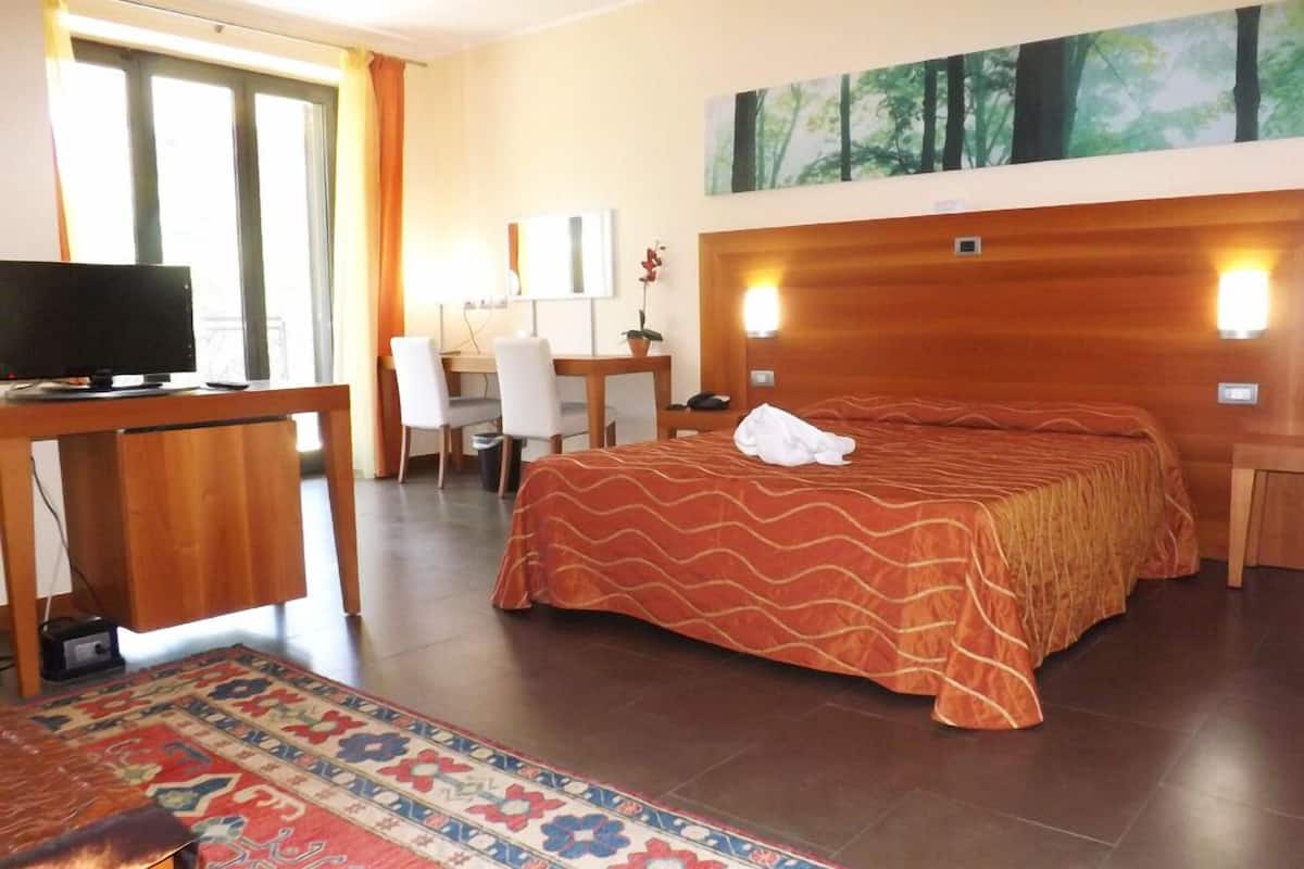 Quarto de hotel com uma cama, uma escrivaninha com um computador, uma televisão e um quadro na parede.
