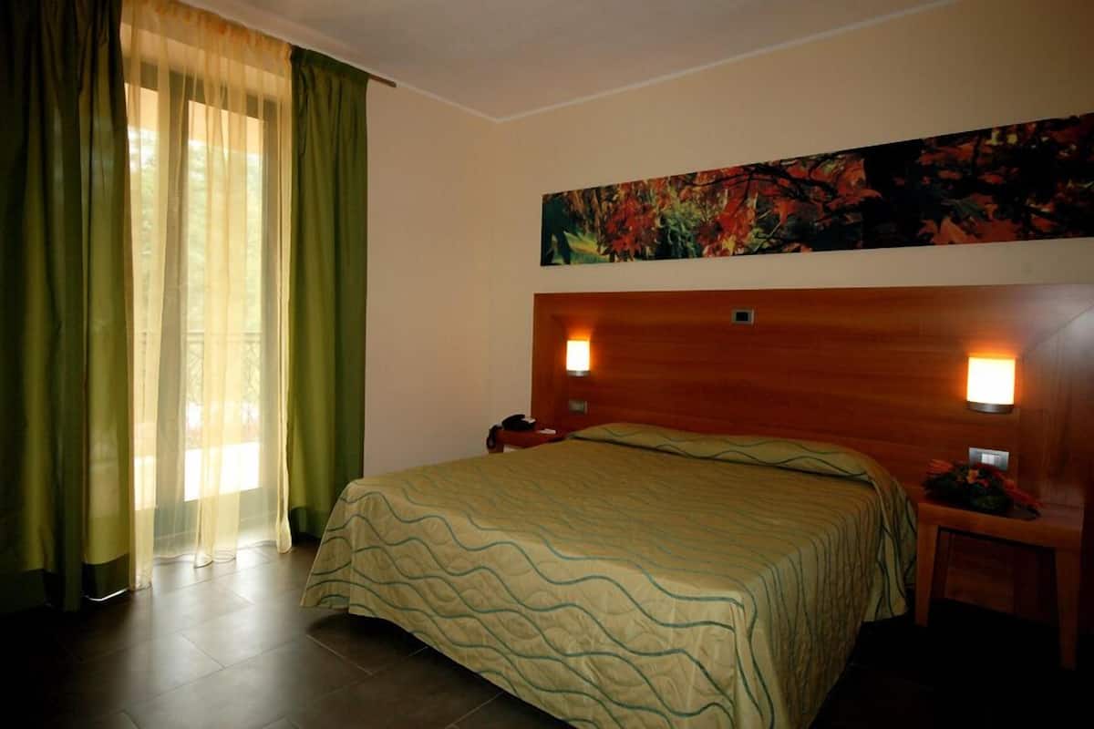 Quarto de hotel com cama, mesa de cabeceira, um quadro e uma janela com cortinas.