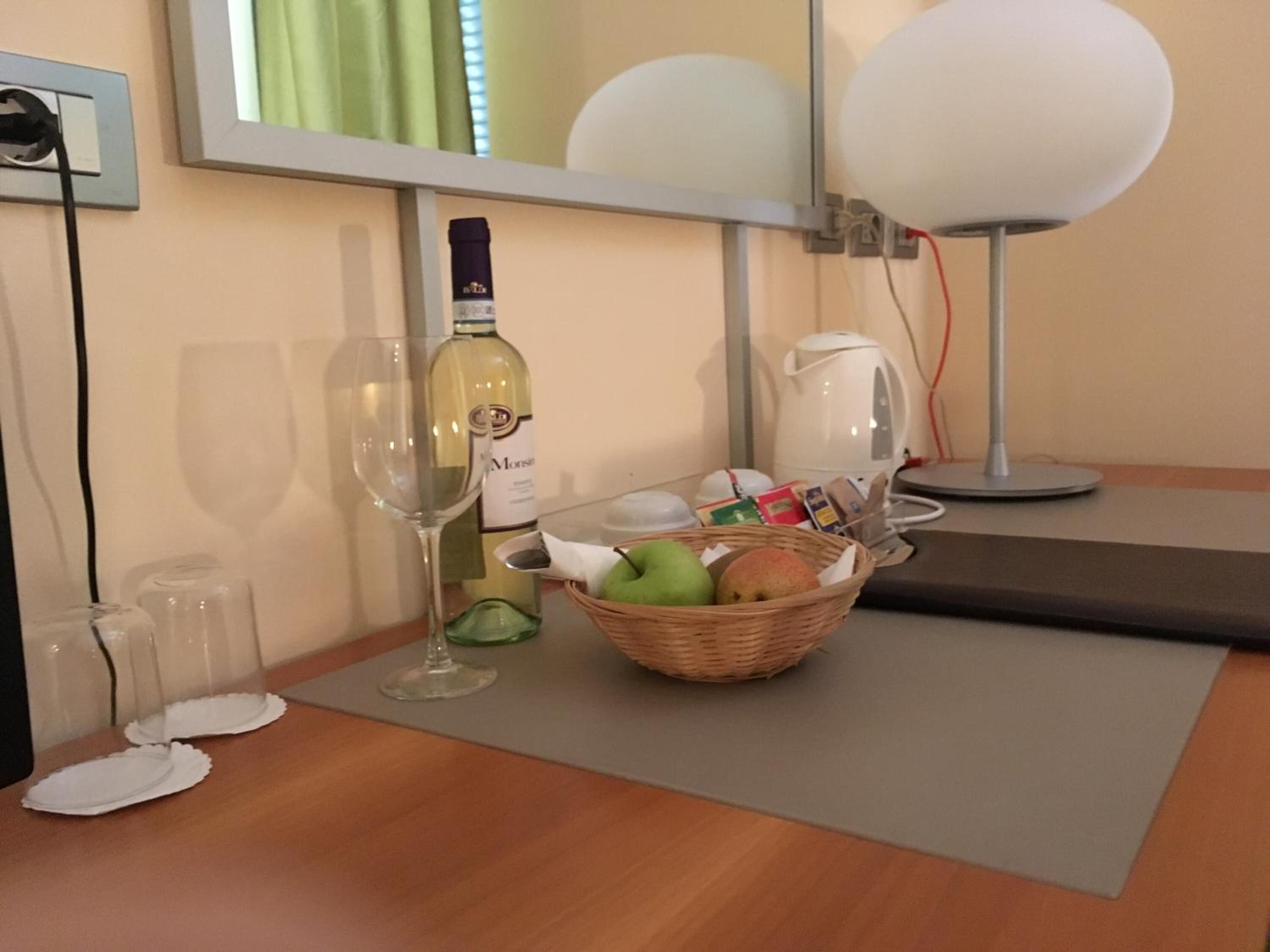 Habitación de hotel con una mesa de comedor, una botella de vino, una cesta de frutas, una cafetera y una lámpara.