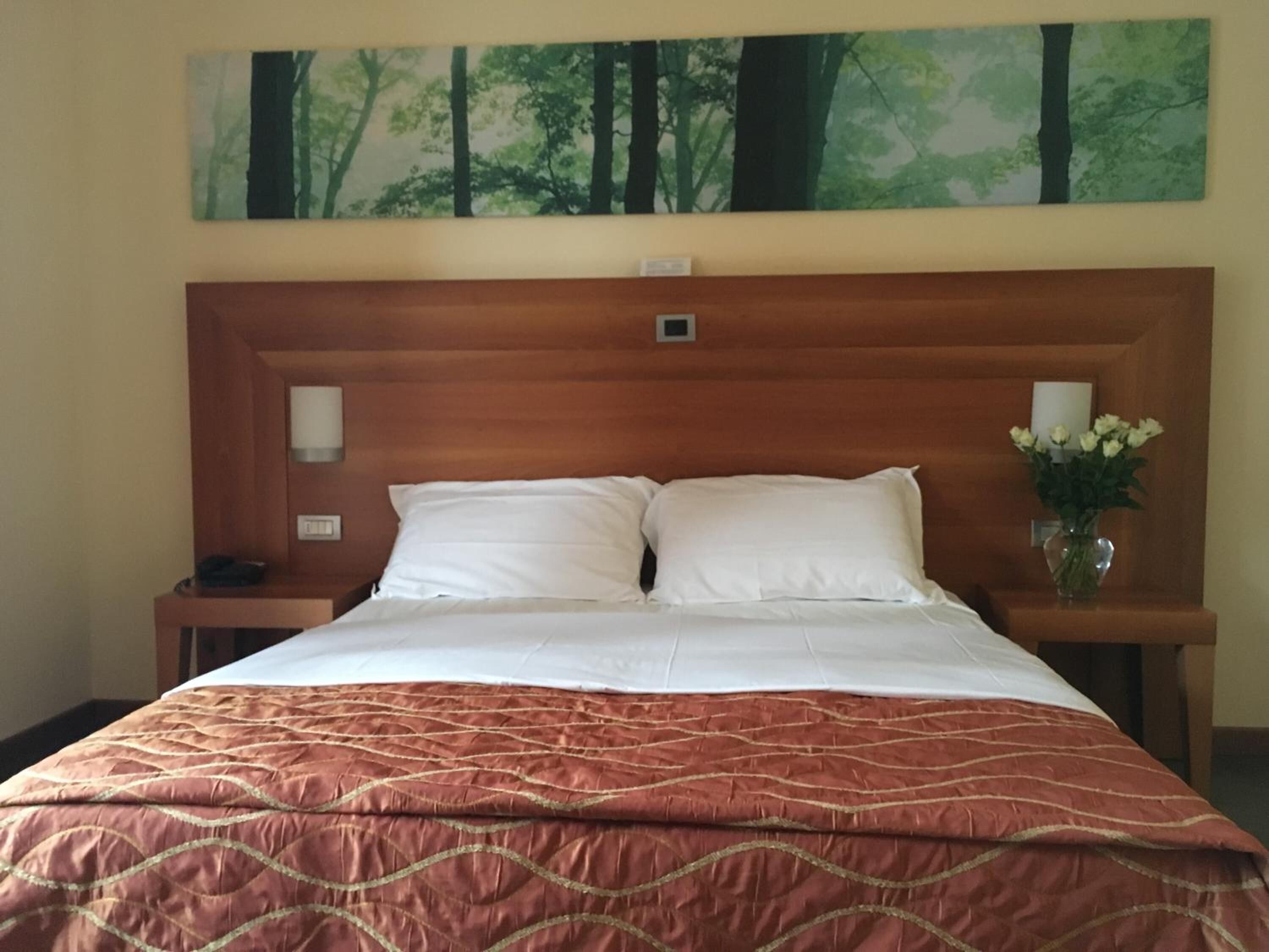 A hotel room with a large bed, two white pillows, a brown quilted bedspread, a nightstand with a phone, a vase with white flowers, and a painting of a forest on the wall.