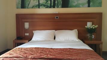 Une chambre d’hôtel avec un grand lit, deux oreillers blancs, une couverture matelassée marron, une table de chevet avec un téléphone, un vase avec des fleurs blanches et un tableau représentant une forêt accroché au mur.