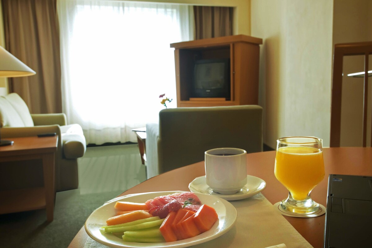 A hotel room with a dining table set with a plate of fruit, a cup of coffee, and a glass of orange juice.