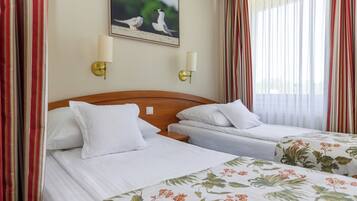 Habitación de hotel con dos camas individuales, cabecero de madera, ropa de cama blanca, colcha con motivos florales, dos lámparas de pared, un cuadro enmarcado de pájaros y cortinas a rayas rojas y blancas.