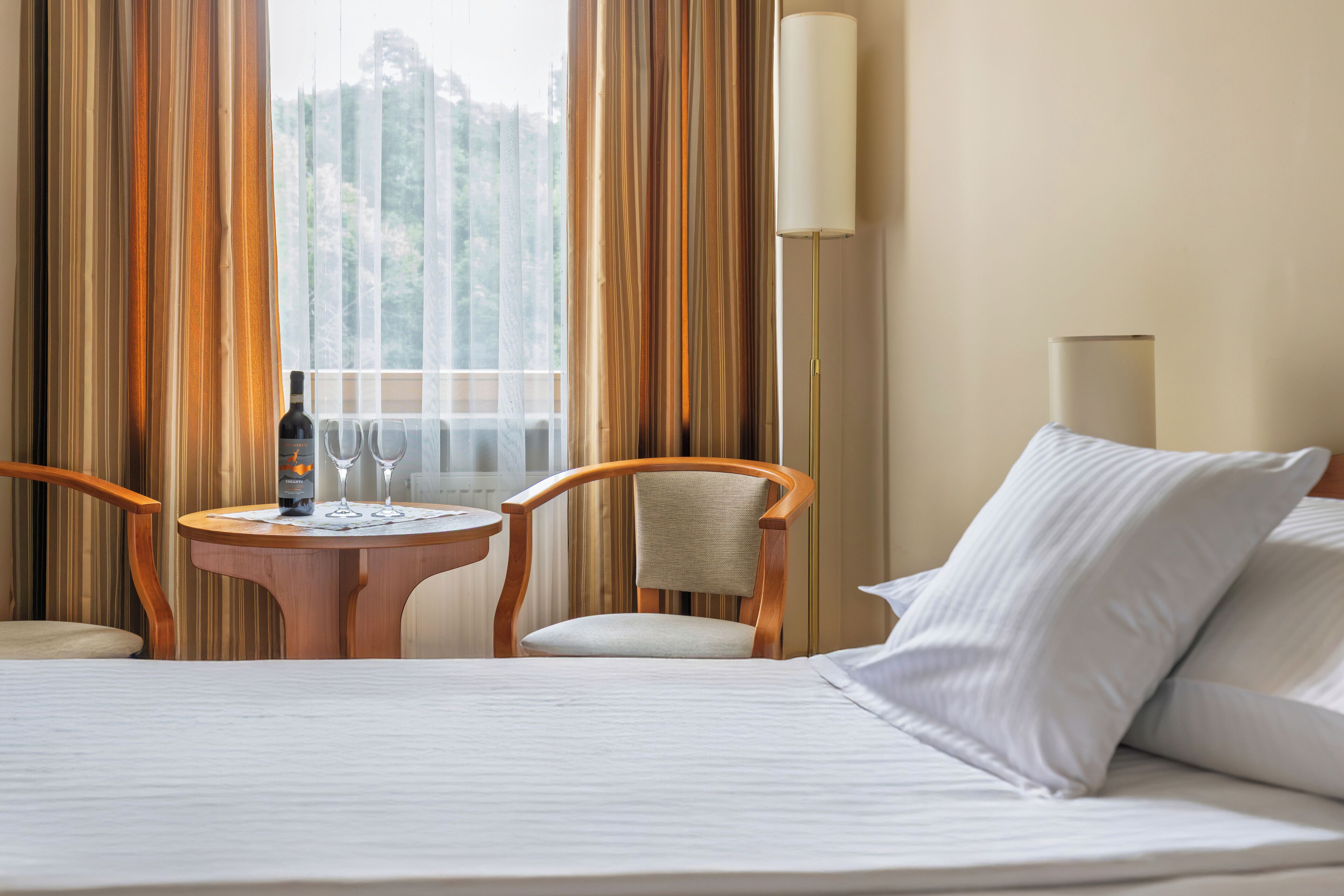 Una camera d'albergo con un letto, due sedie, un tavolino con una bottiglia e dei bicchieri, e una lampada.