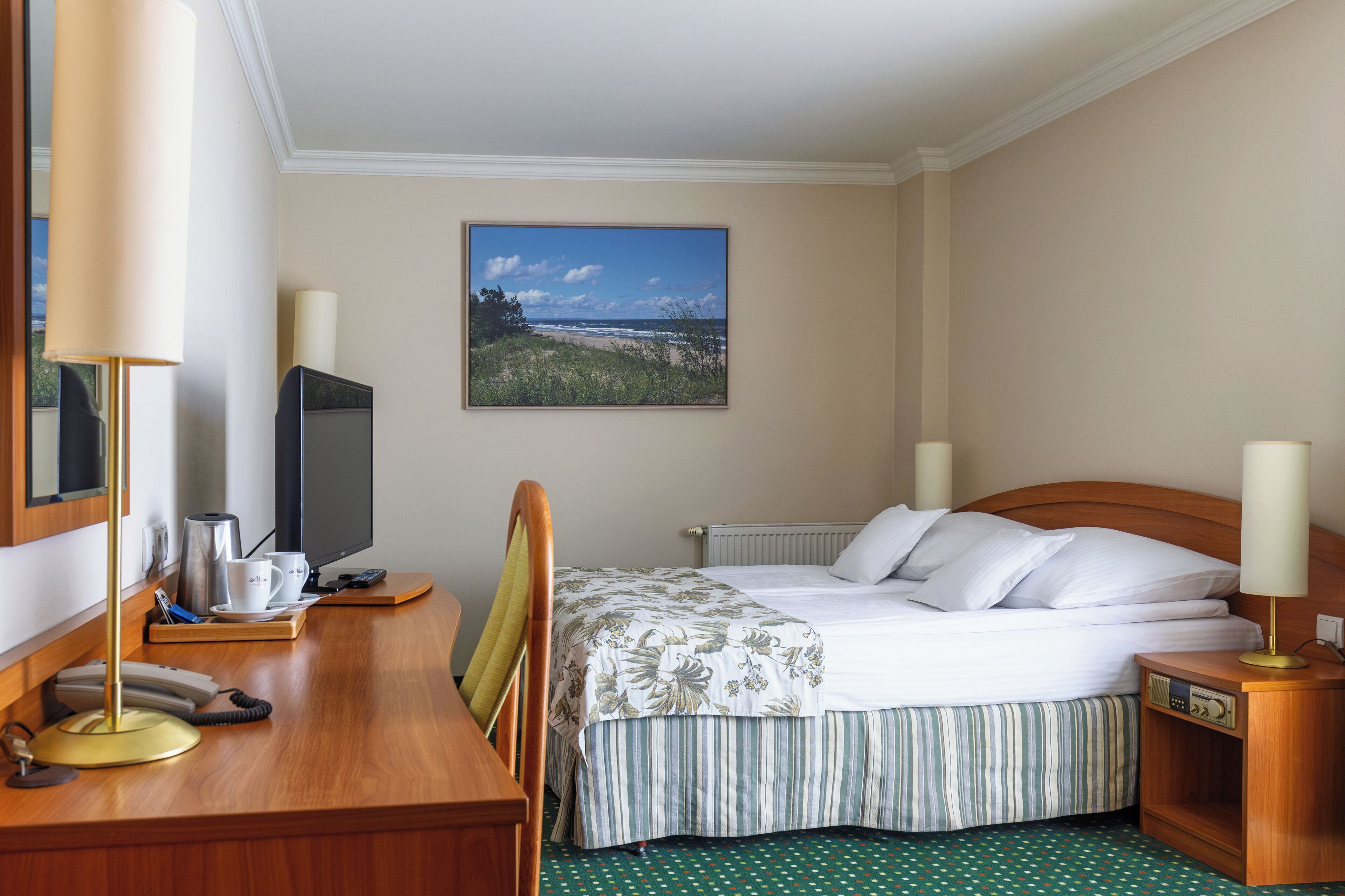 A hotel room with a bed, a desk with a computer, a lamp, and a painting of a beach scene.