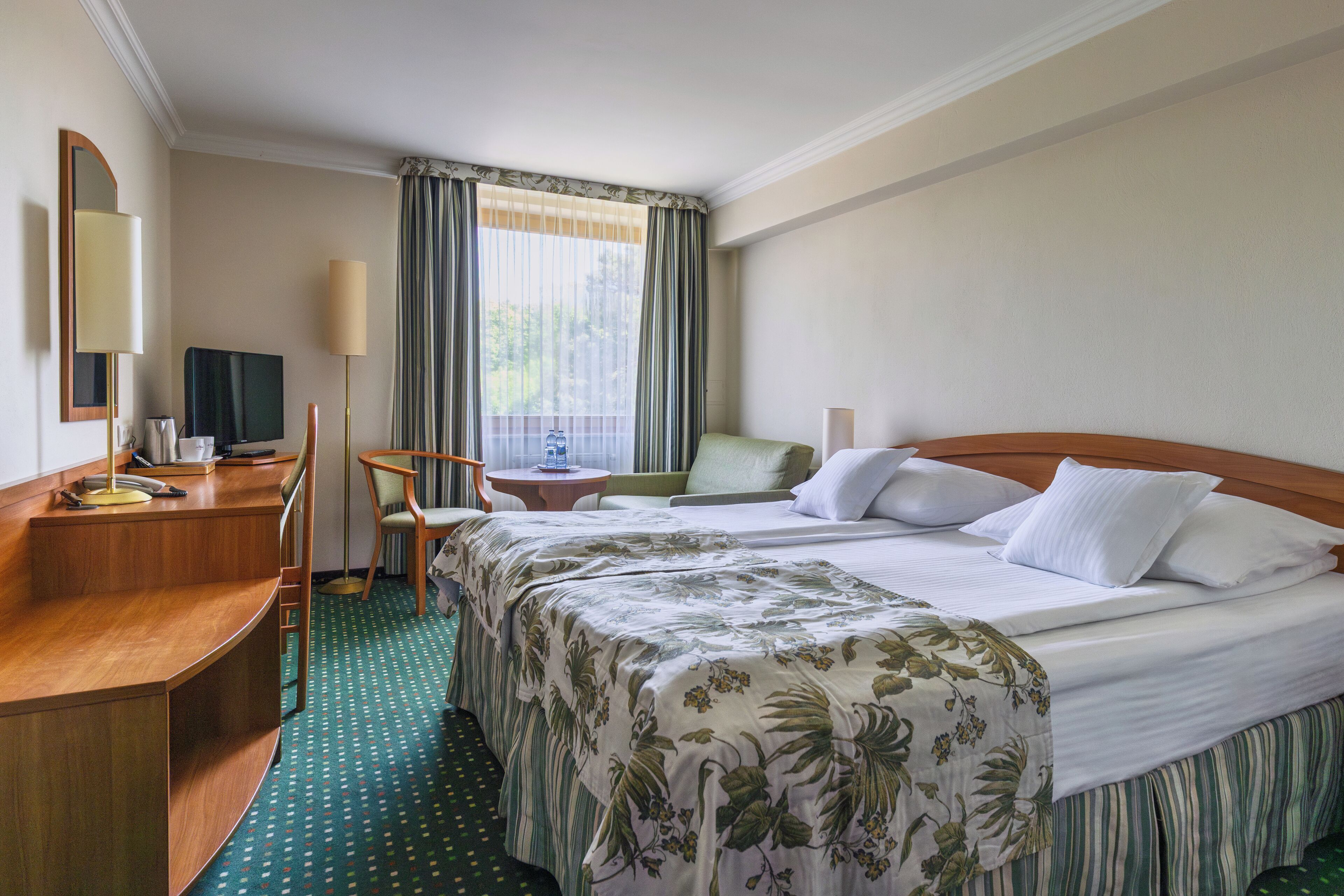 Habitación de hotel con una cama grande, un escritorio, una silla, un televisor y una ventana con cortinas.