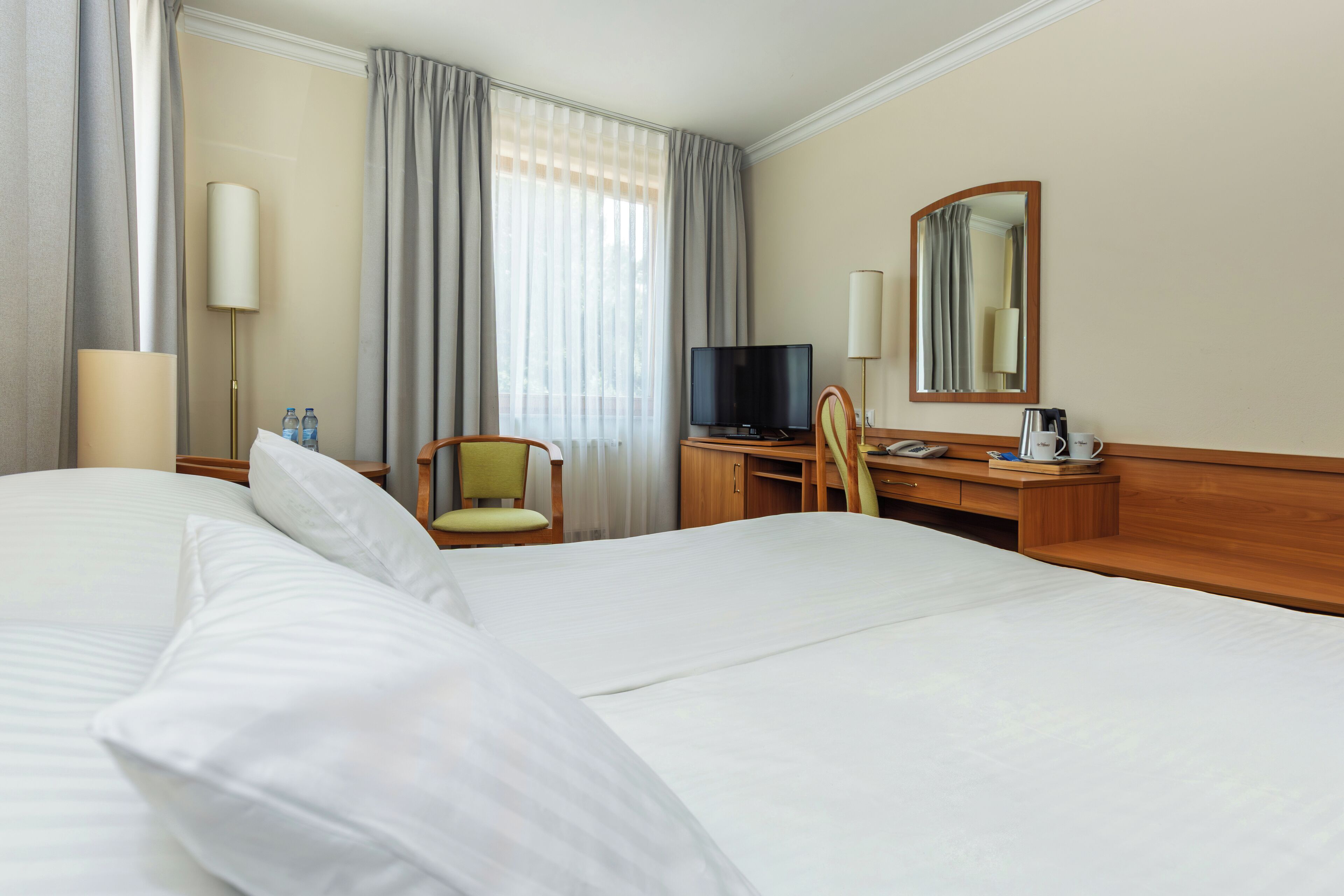 A hotel room with a large bed, a desk with a chair, a television, a mirror, and a window with curtains.