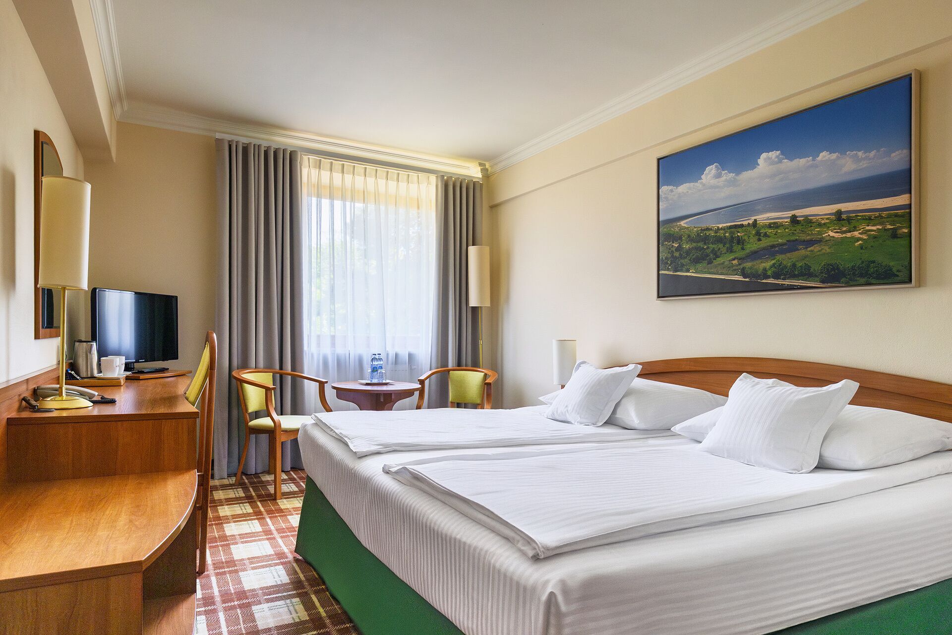 Hotelkamer met twee bedden, een bureau met een computer, een stoel, een televisie en een landschapsdecoratie.