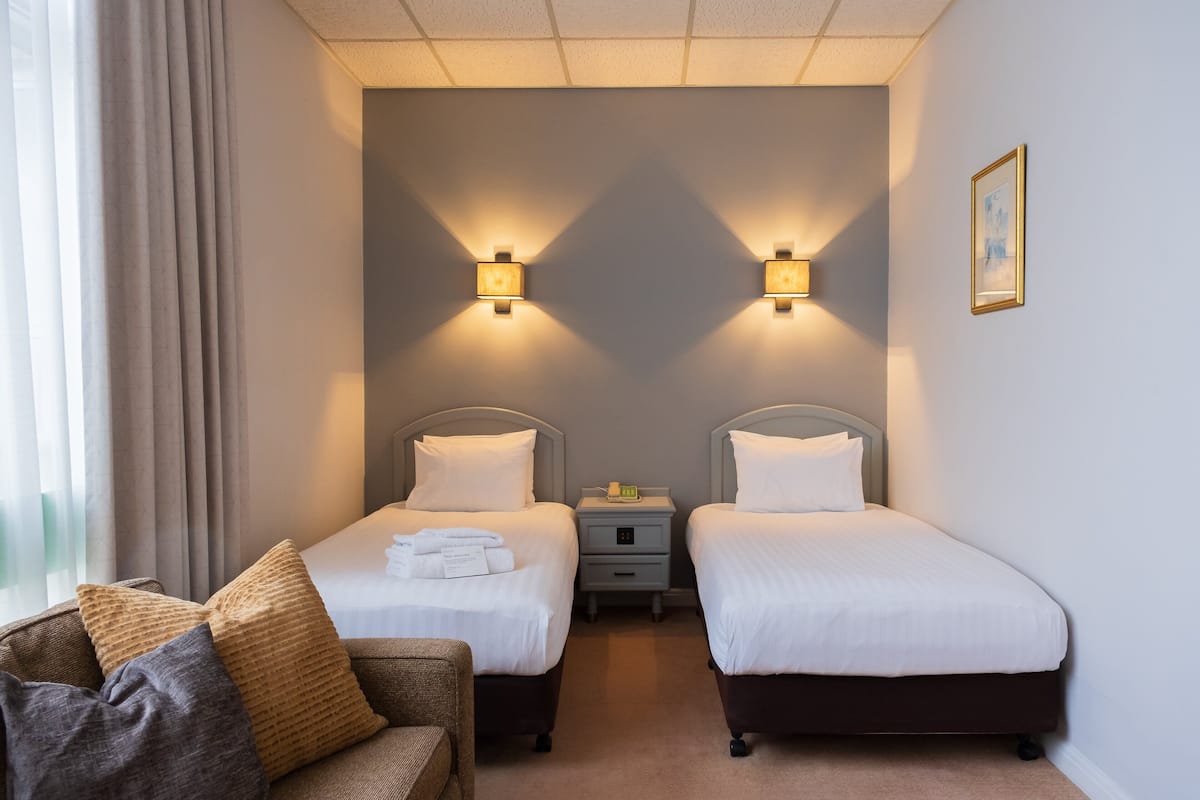 A hotel room with two single beds, a small bedside table with a lamp, a sofa with cushions, and a framed picture on the wall.