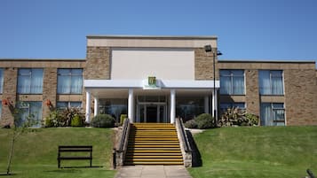 A Holiday Inn with a covered entrance, a staircase leading up to the entrance, and a grassy area with a bench.