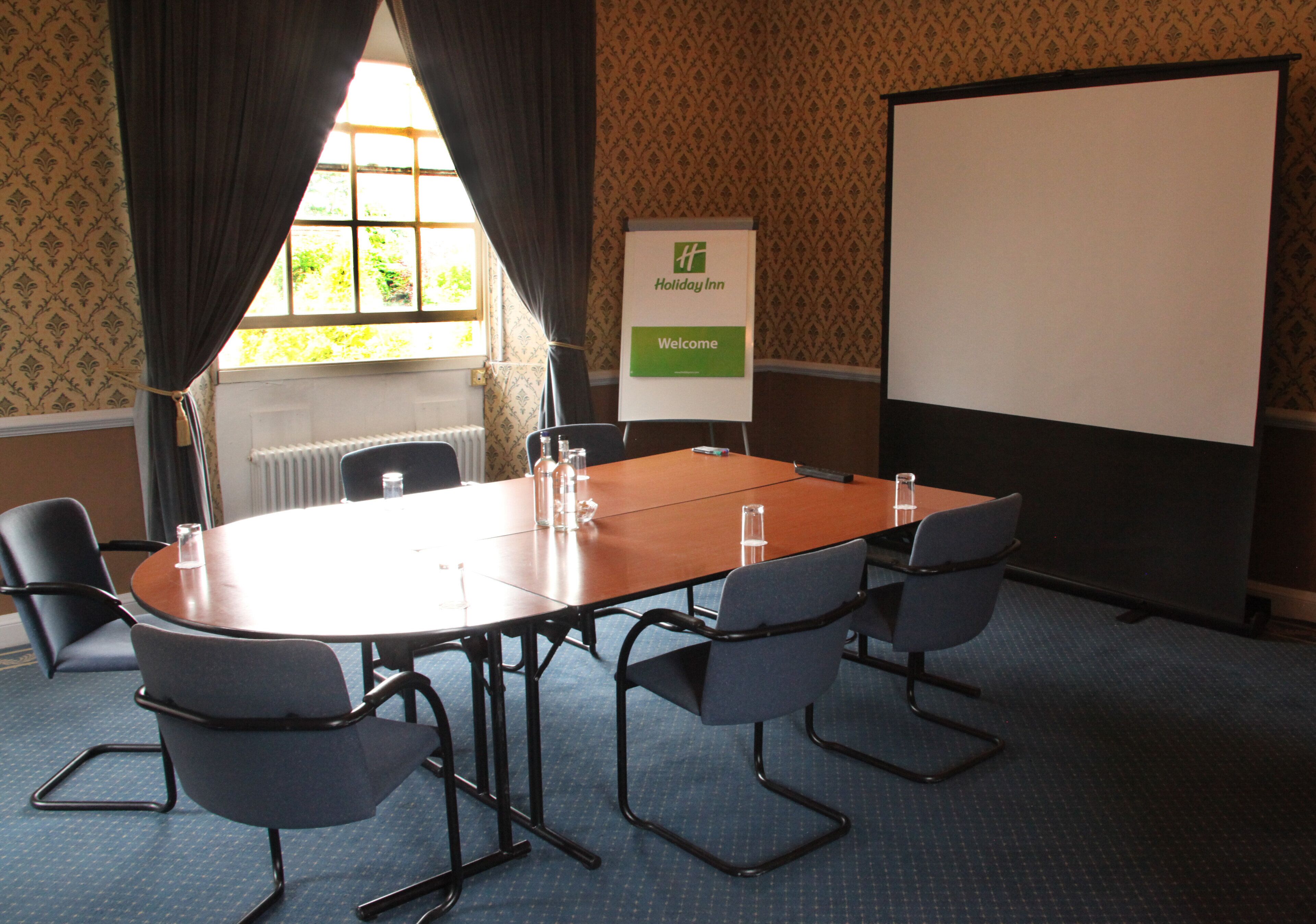 Una sala de conferencias con una mesa grande, seis sillas, una pantalla de proyector y un letrero de Holiday Inn.