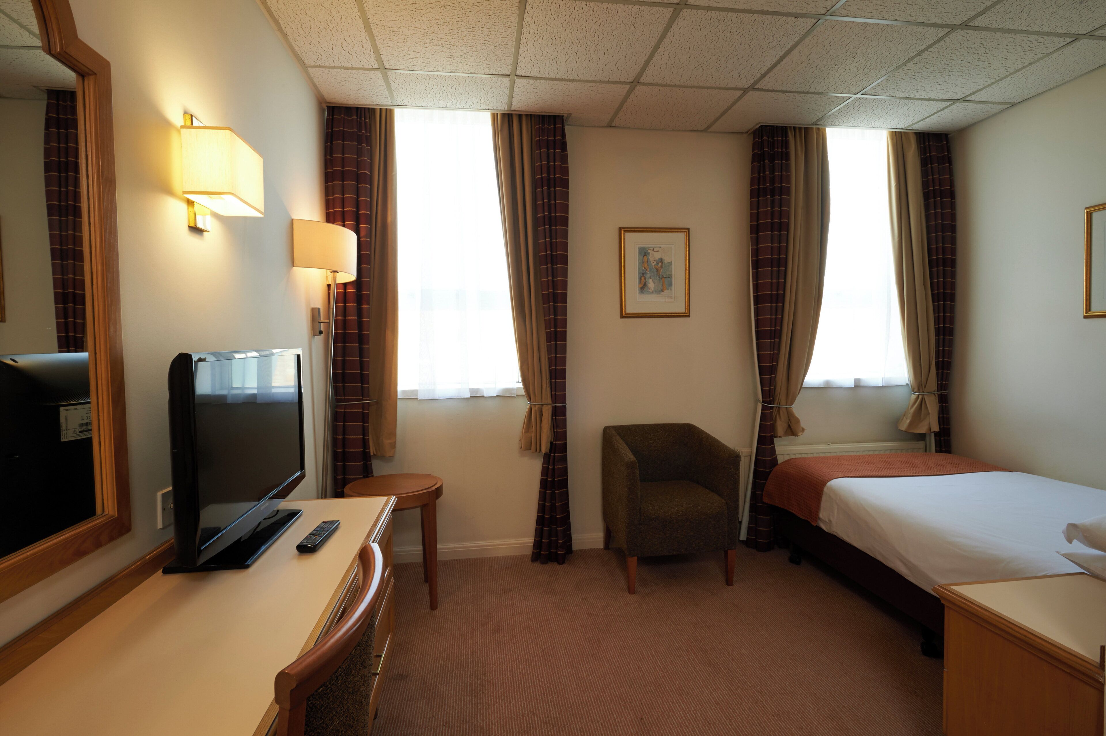 A hotel room with a bed, a desk with a television, a chair, and a window with curtains.