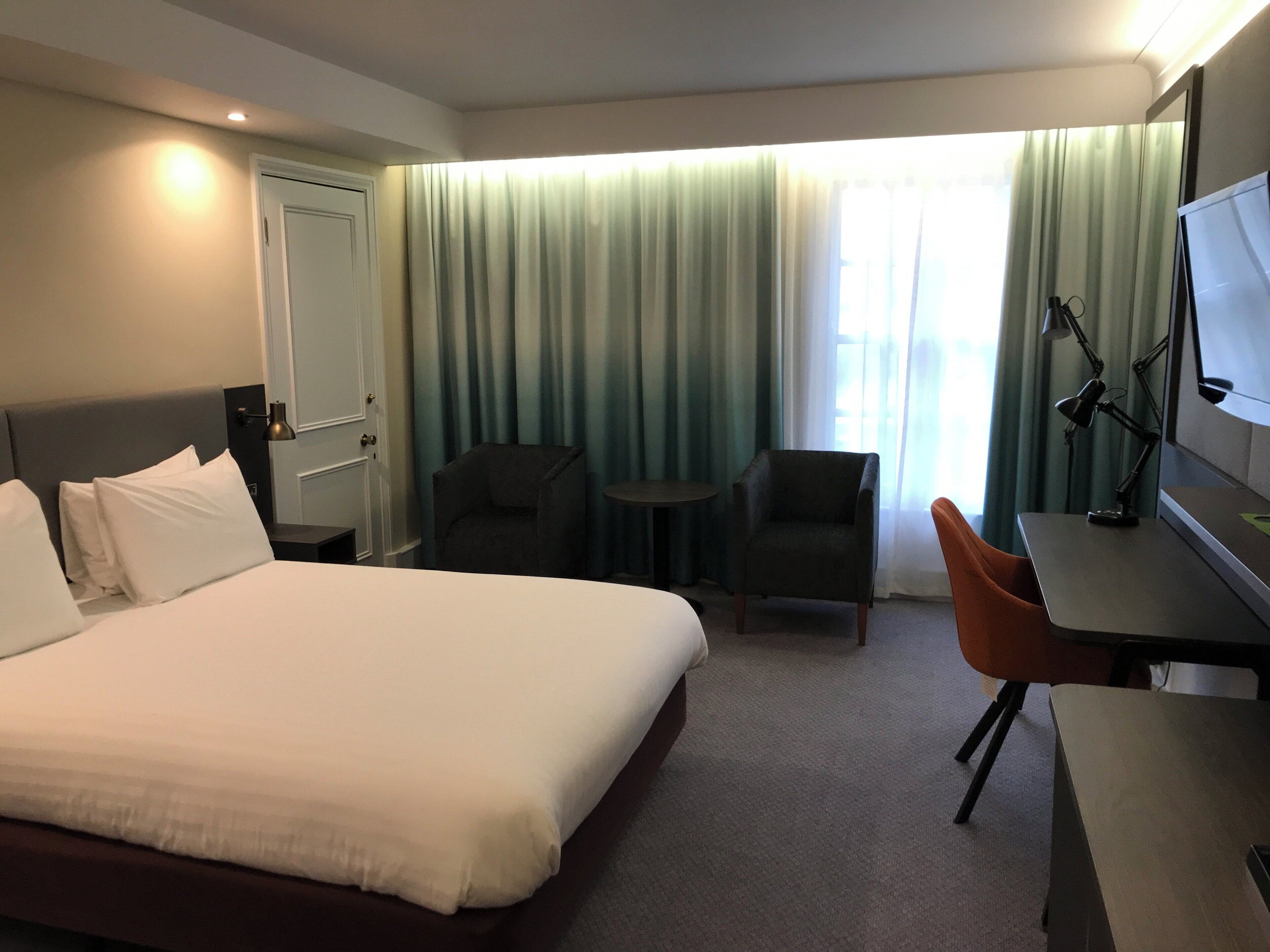 Quarto de hotel com cama, duas poltronas, escrivaninha, televisão e janela com cortinas.