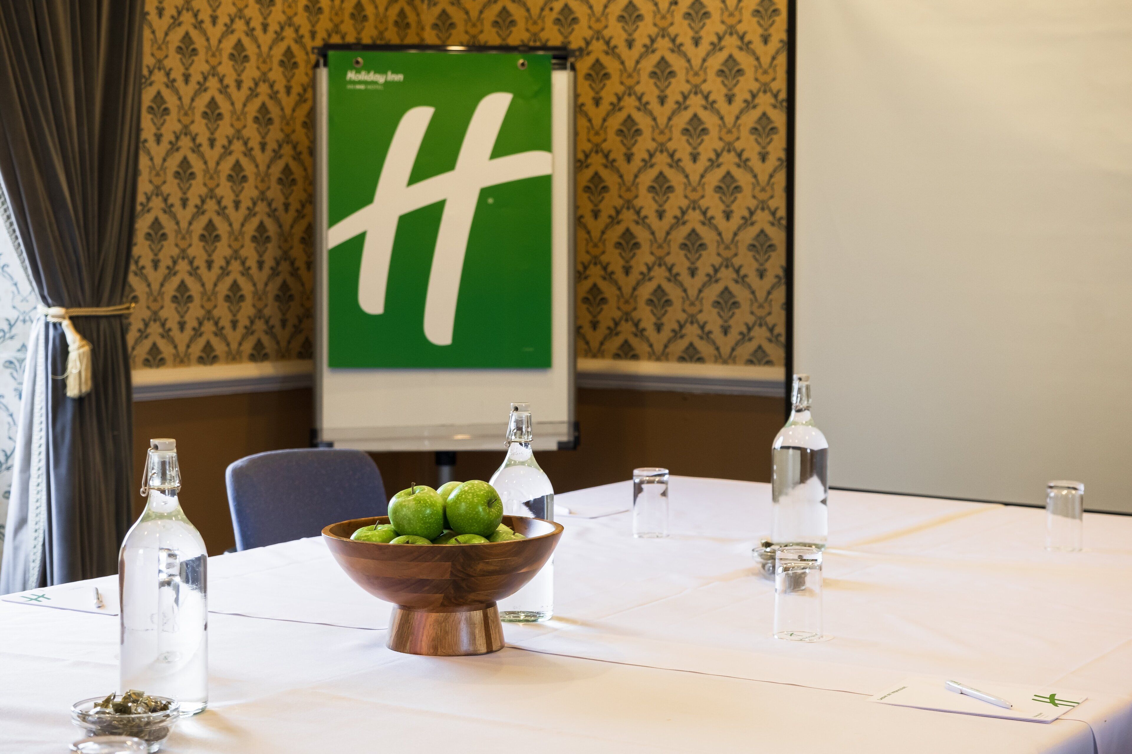 Uma sala de conferências com uma mesa branca, uma tigela de madeira com maçãs verdes, garrafas de água transparentes e uma tela de projetor com um logotipo verde.