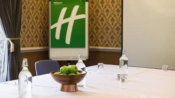 Uma sala de conferências com uma mesa branca, uma tigela de madeira com maçãs verdes, garrafas de água transparentes e uma tela de projetor com um logotipo verde.