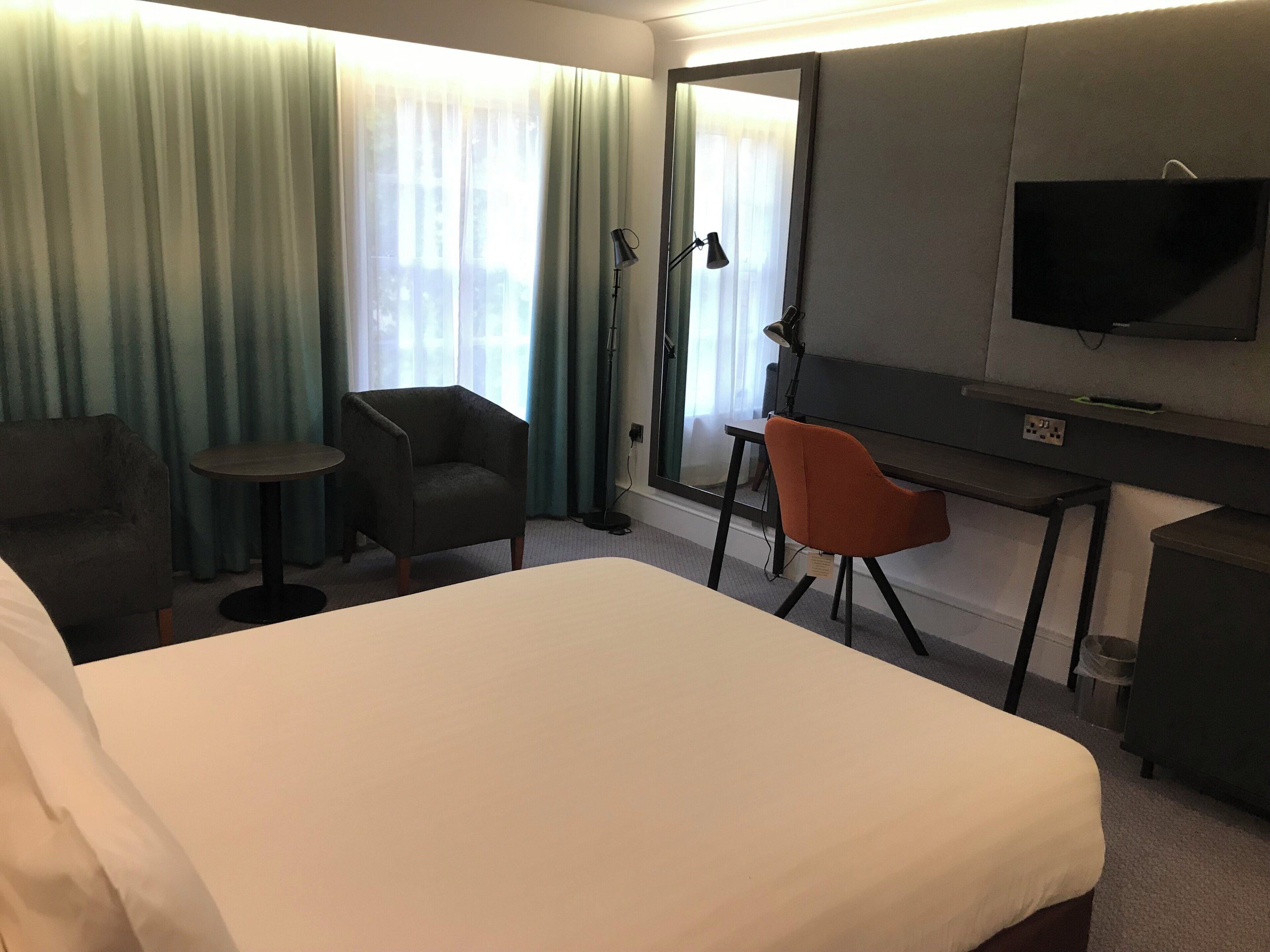 Una habitación de hotel con una cama, dos sillas, un escritorio, un televisor y un ventanal con cortinas.
