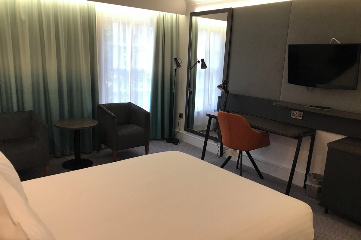 A hotel room with a large bed, two armchairs, a desk, a television, and a window with curtains.