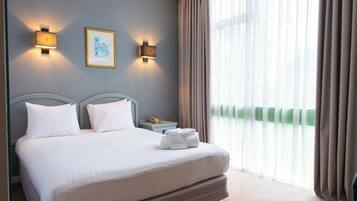 A hotel room with a bed, two wall-mounted lights, a framed picture, a bedside table with a phone, and a window with curtains.