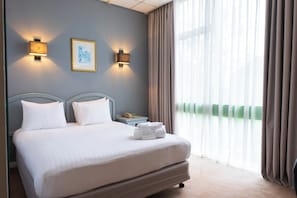 A hotel room with a bed, two wall-mounted lights, a framed picture, a bedside table with a phone, and a window with curtains.