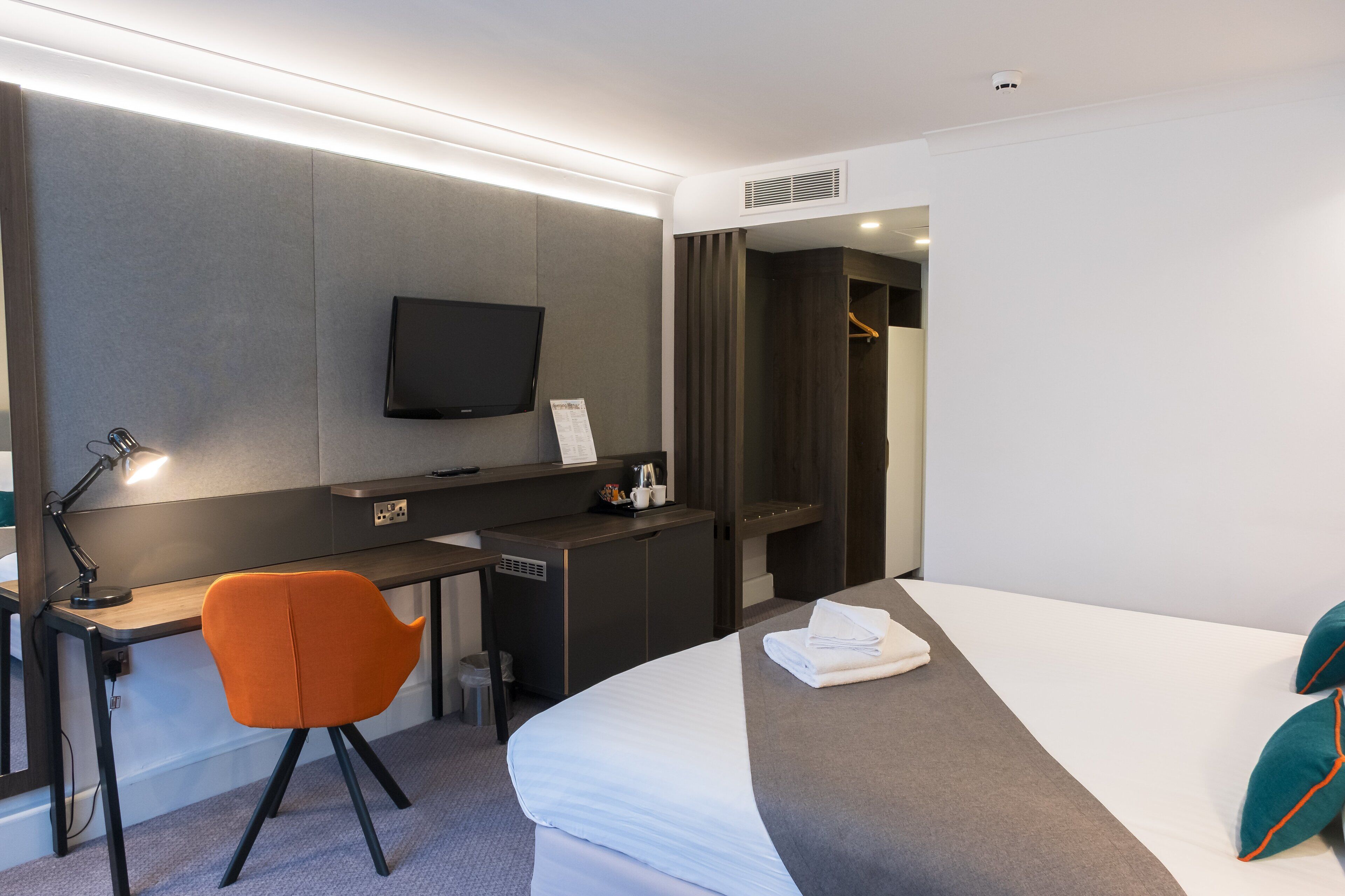 A modern hotel room with a desk, chair, bed, TV, and a walk-in closet.