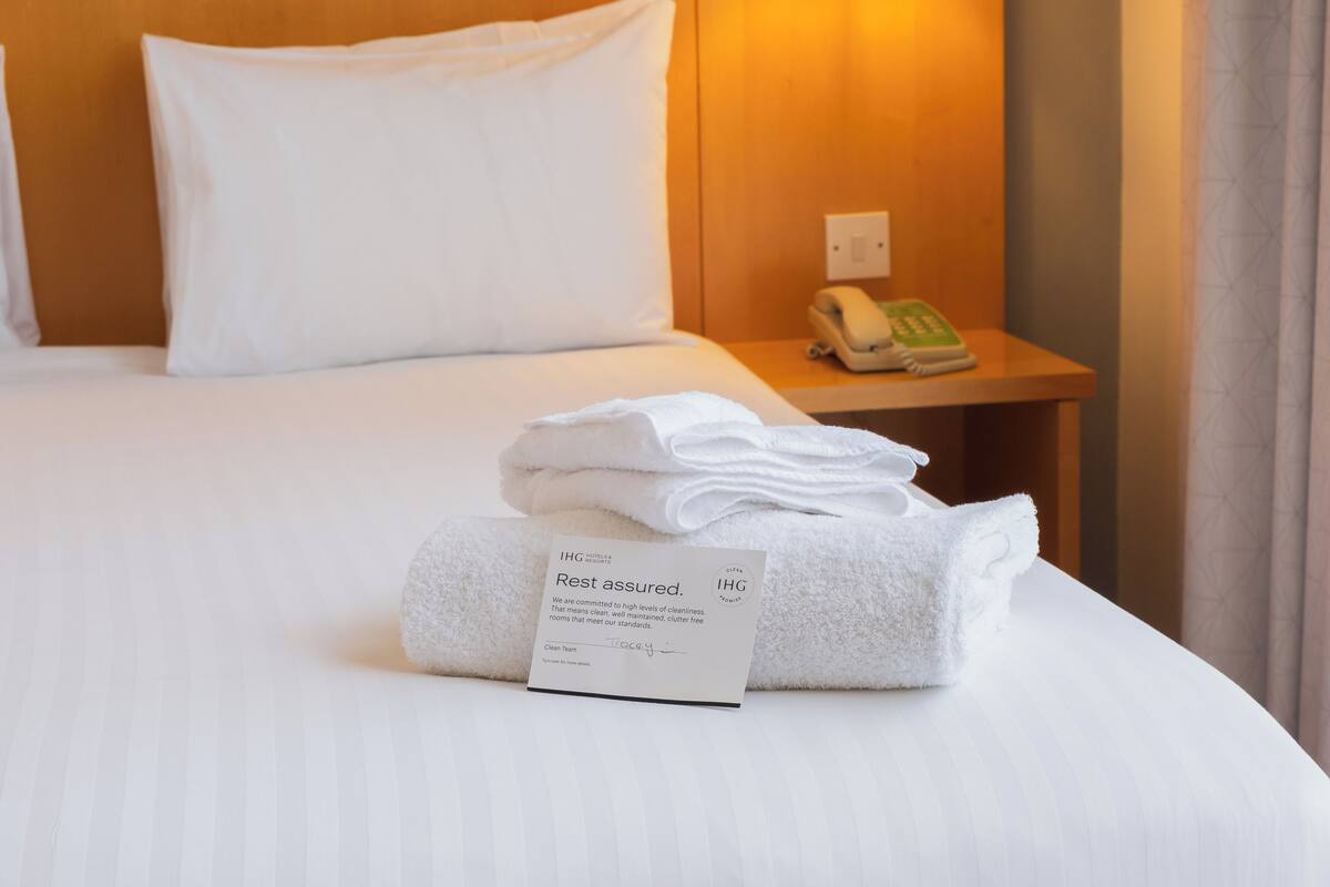 A hotel room with a neatly made bed, a folded towel, a sign indicating room standards, and a bedside phone.