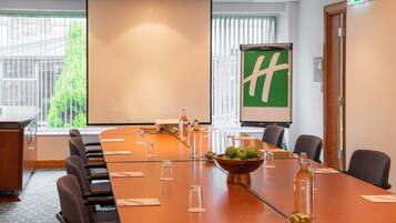 A conference room with a long table, chairs, a projector screen, and a green logo on the wall.