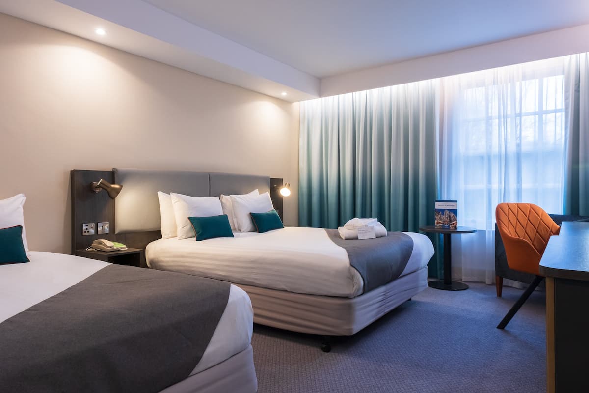A hotel room with two beds, a desk, a chair, and a small table with a book.