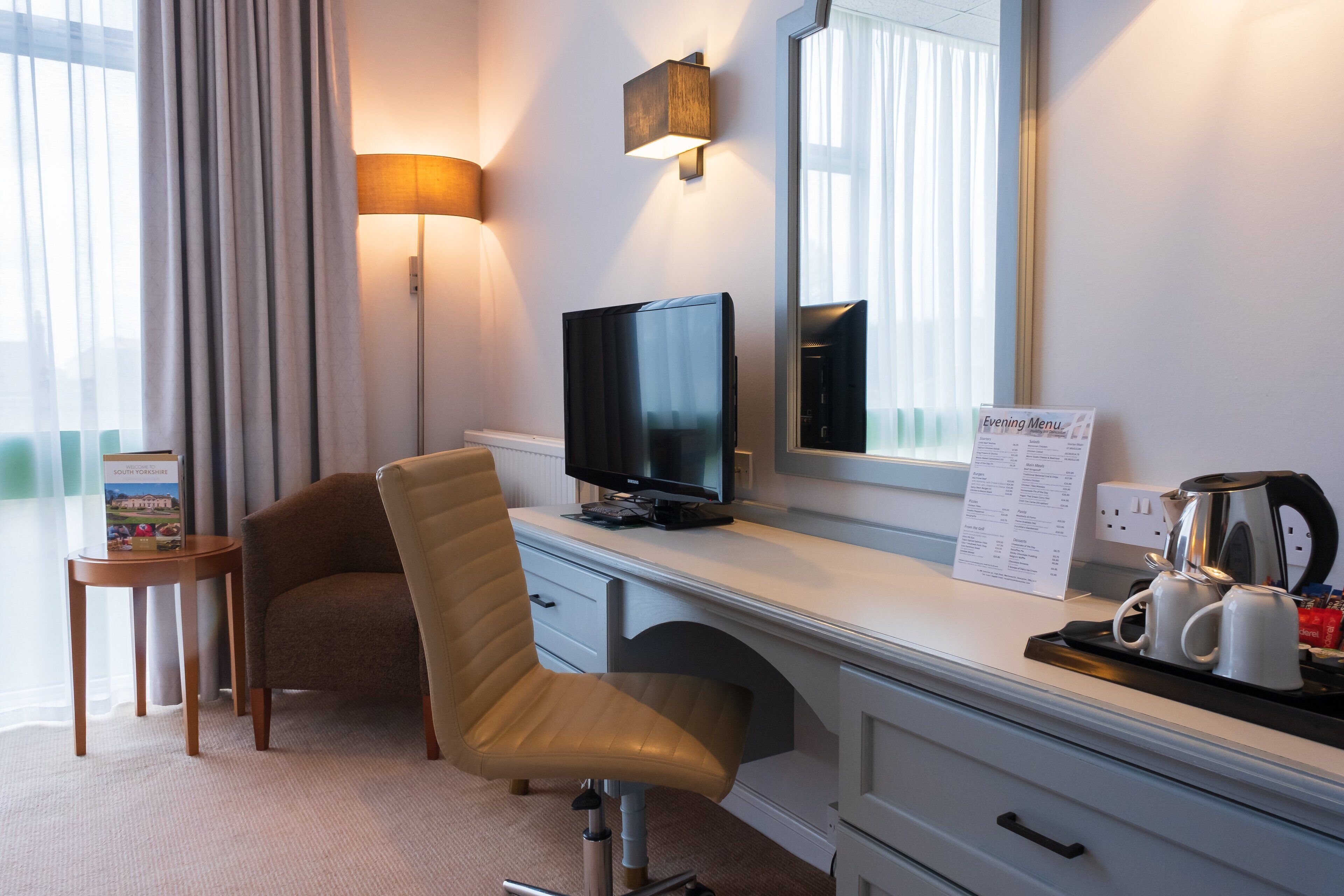 A hotel room with a desk, a television, a chair, a coffee maker, and a menu sign.
