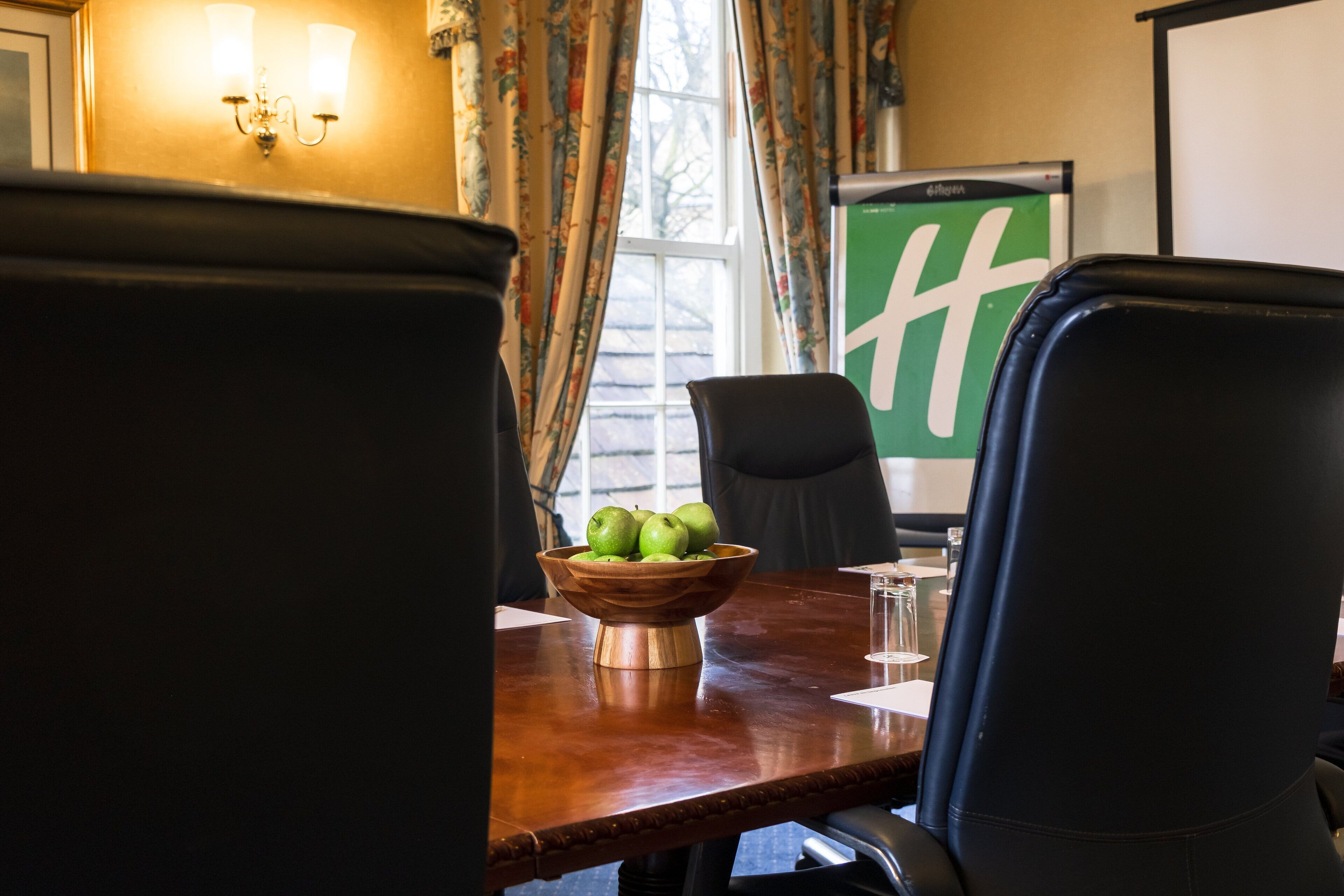 Un salón de conferencias con una mesa de madera, un cuenco de manzanas verdes, dos sillas negras y una pantalla de proyector con un logotipo verde.