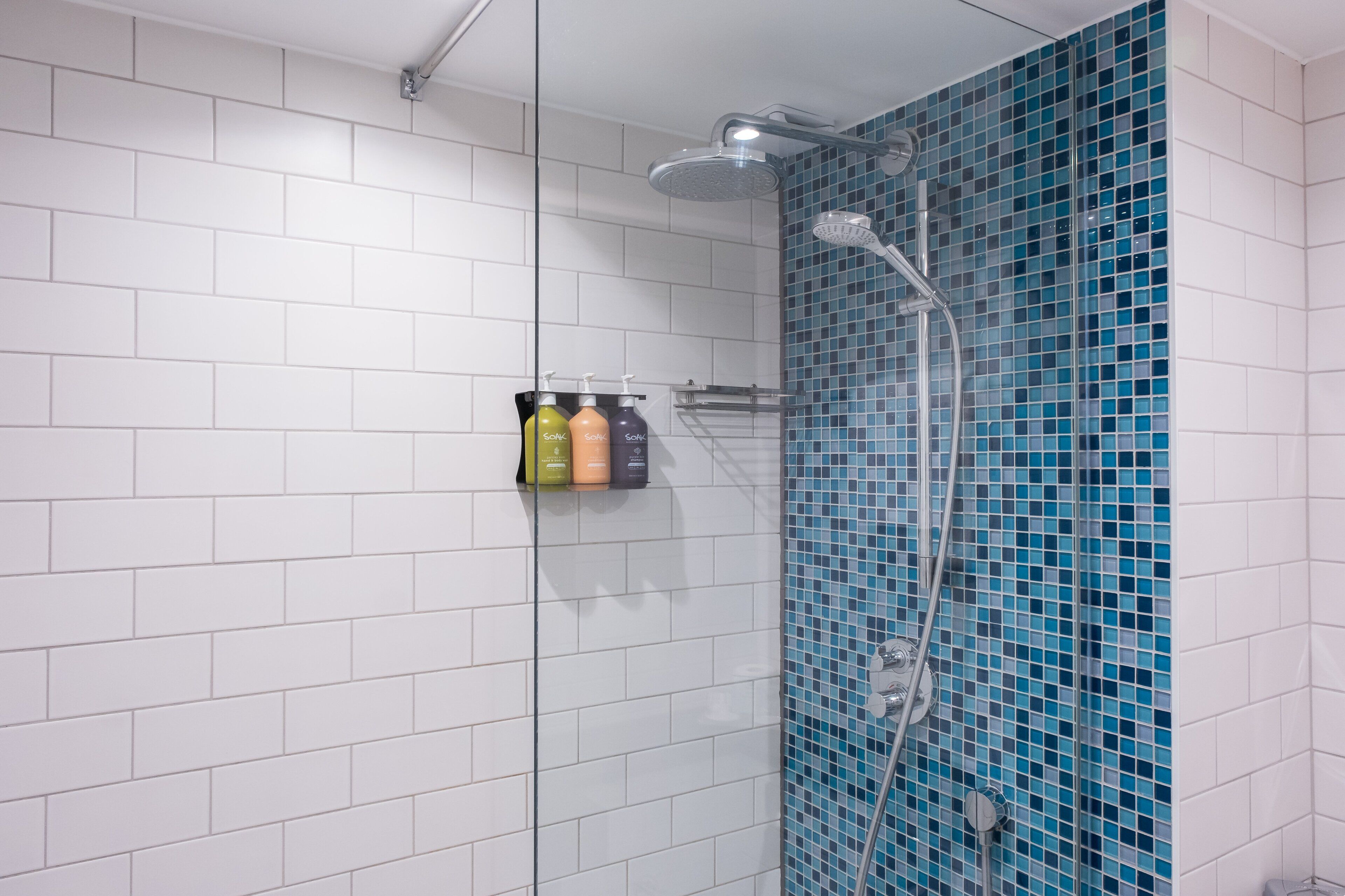 Una ducha con pared de mosaico azul, suelo de baldosas blancas y estante con tres botellas de champú.