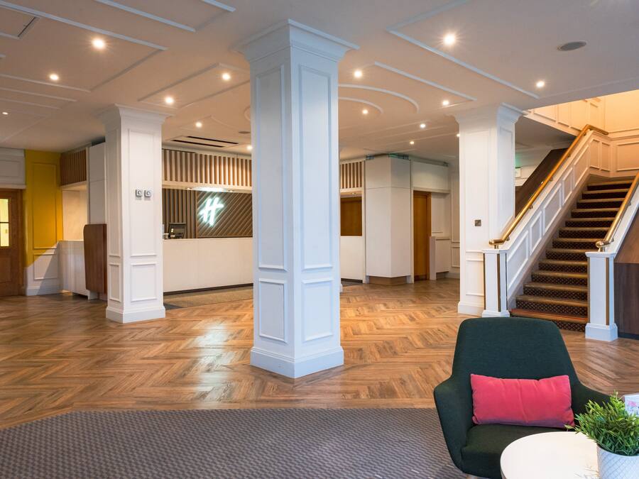 A spacious lobby with a wooden floor, white columns, a staircase, and a green armchair with a red pillow.