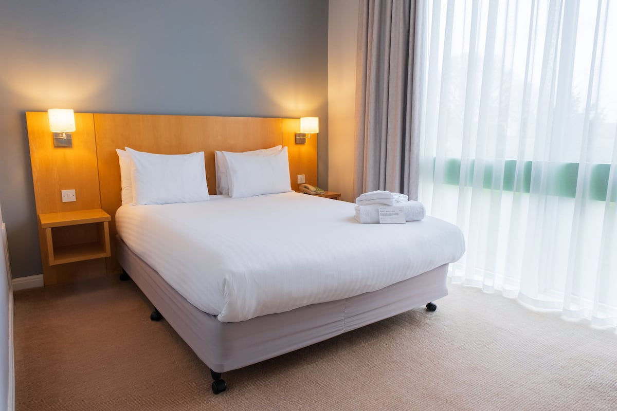 A hotel room with a large bed, two bedside lamps, a nightstand, and a window with sheer curtains.