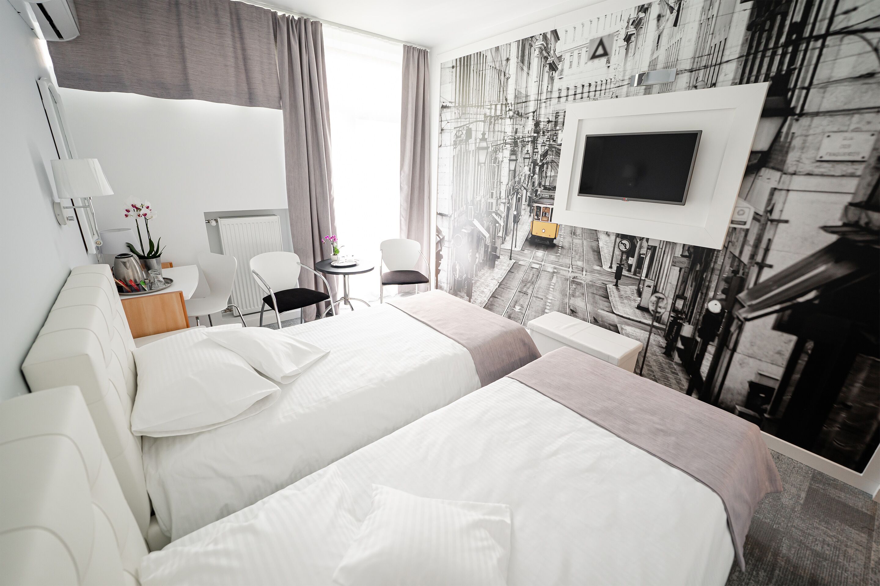 A hotel room with two beds, a TV mounted on the wall, a small table with a lamp, and a view of a city street through the window.