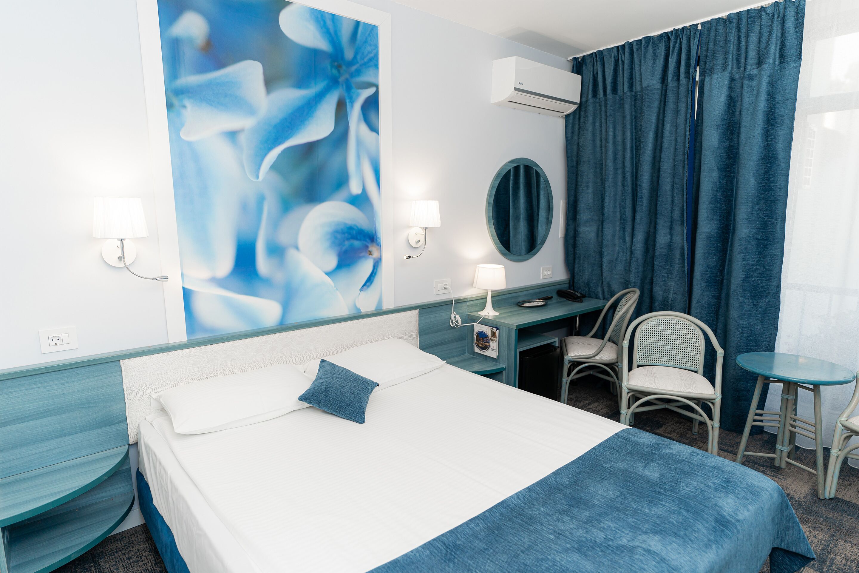 Una habitación de hotel con cama, escritorio, dos sillas, un espejo redondo y un cuadro floral en la pared.