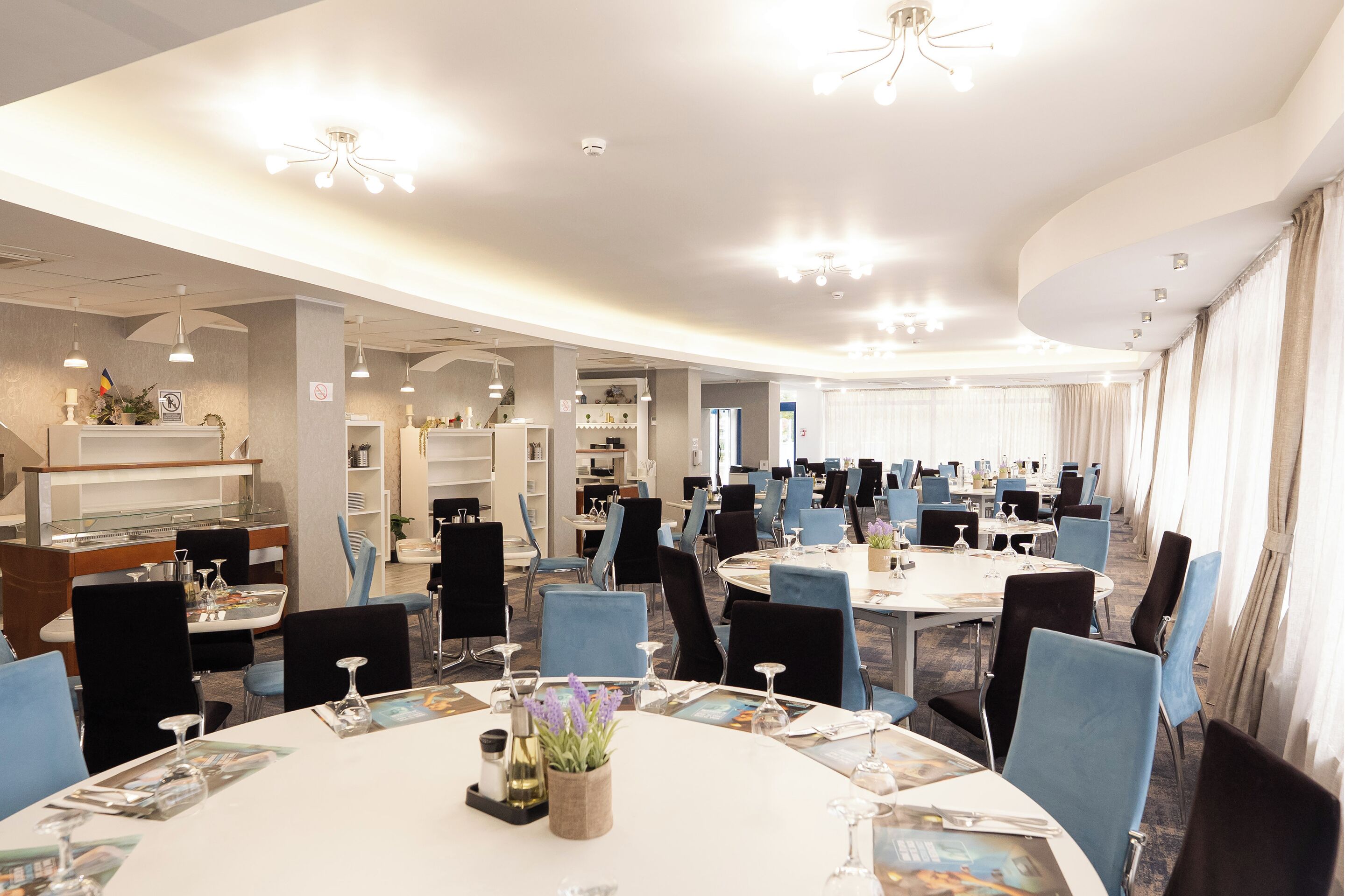 Un amplio comedor de hotel con varias mesas redondas, sillas azules y modernas lámparas.