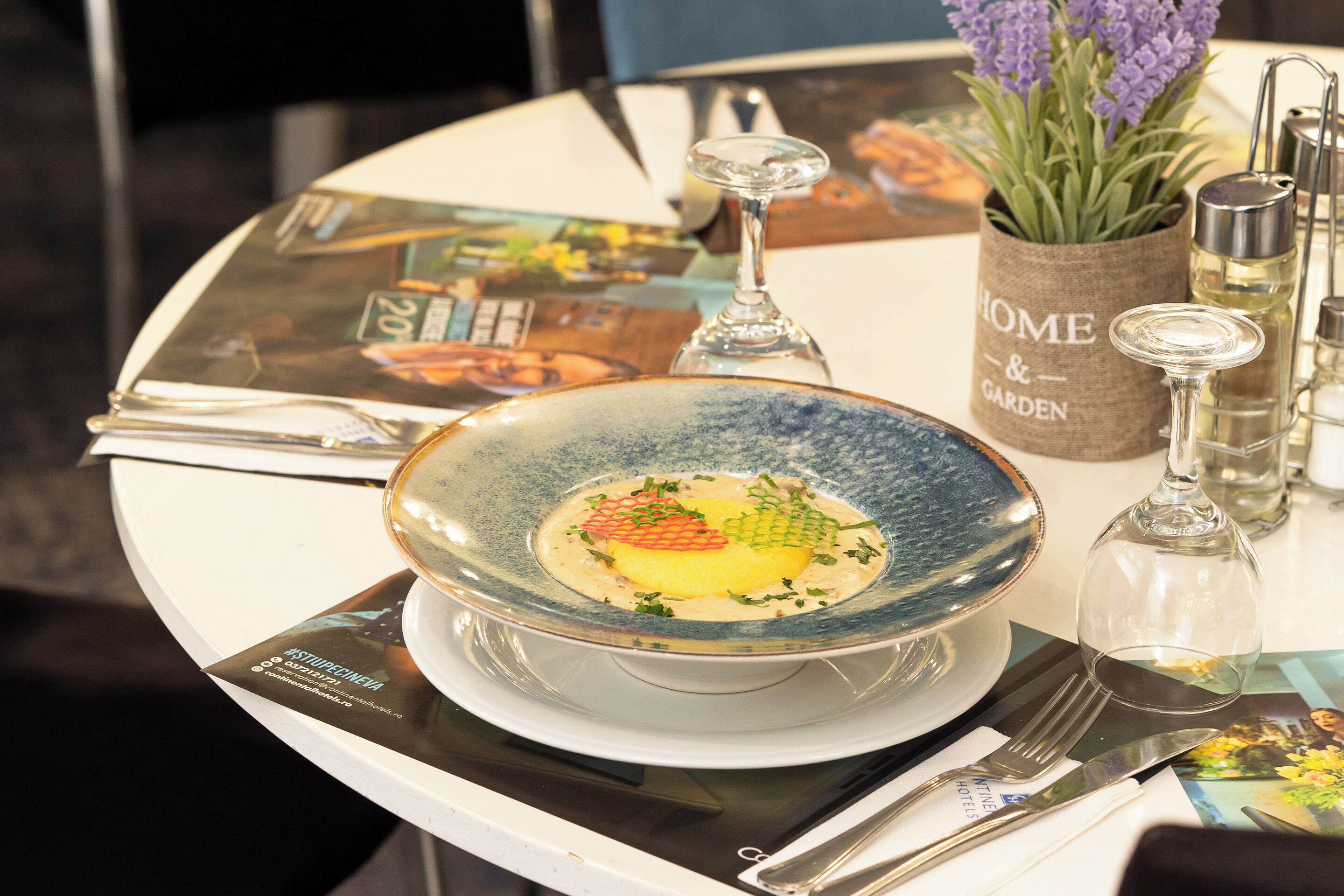 A dining table with a blue ceramic bowl containing a dish, a wine glass, a small vase with lavender, and a menu.