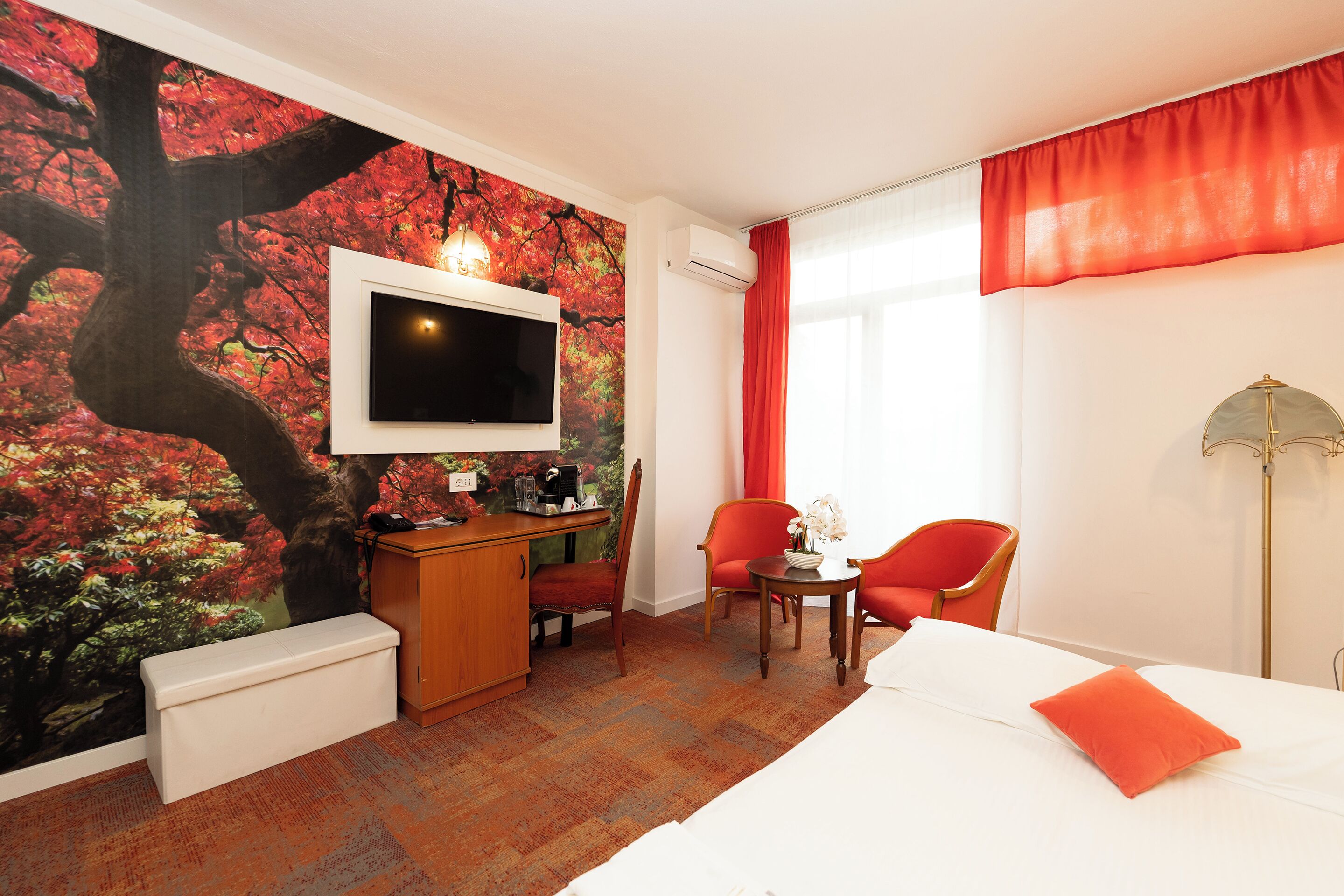 A hotel room with a large tree mural, a flat-screen TV, a desk with a chair, a small table with a vase, and a bed with an orange pillow.