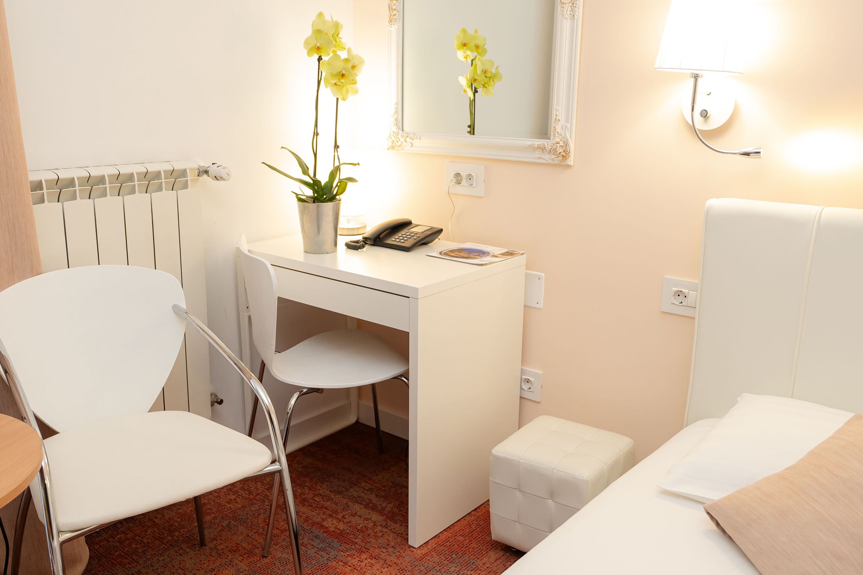 Une chambre d’hôtel avec un bureau blanc, une chaise, un petit pouf blanc, un lit avec une literie blanche, un miroir, une lampe et une plante en pot avec des fleurs jaunes.
