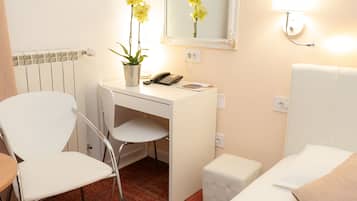 A hotel room with a white desk, a chair, a small white ottoman, a bed with white bedding, a mirror, a lamp, and a potted plant with yellow flowers.