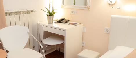 Une chambre d’hôtel avec un bureau blanc, une chaise, un petit pouf blanc, un lit avec une literie blanche, un miroir, une lampe et une plante en pot avec des fleurs jaunes.