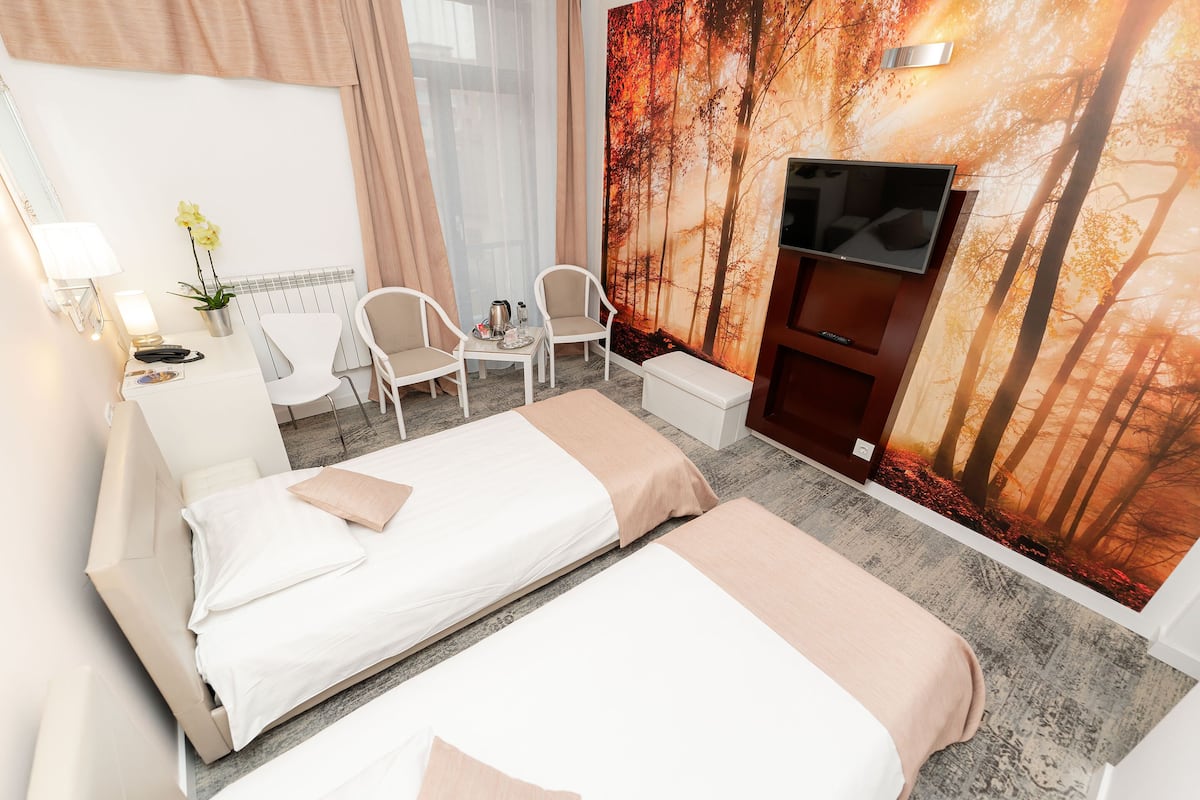 A hotel room with two beds, a TV, a desk with chairs, and a large forest-themed wall mural.