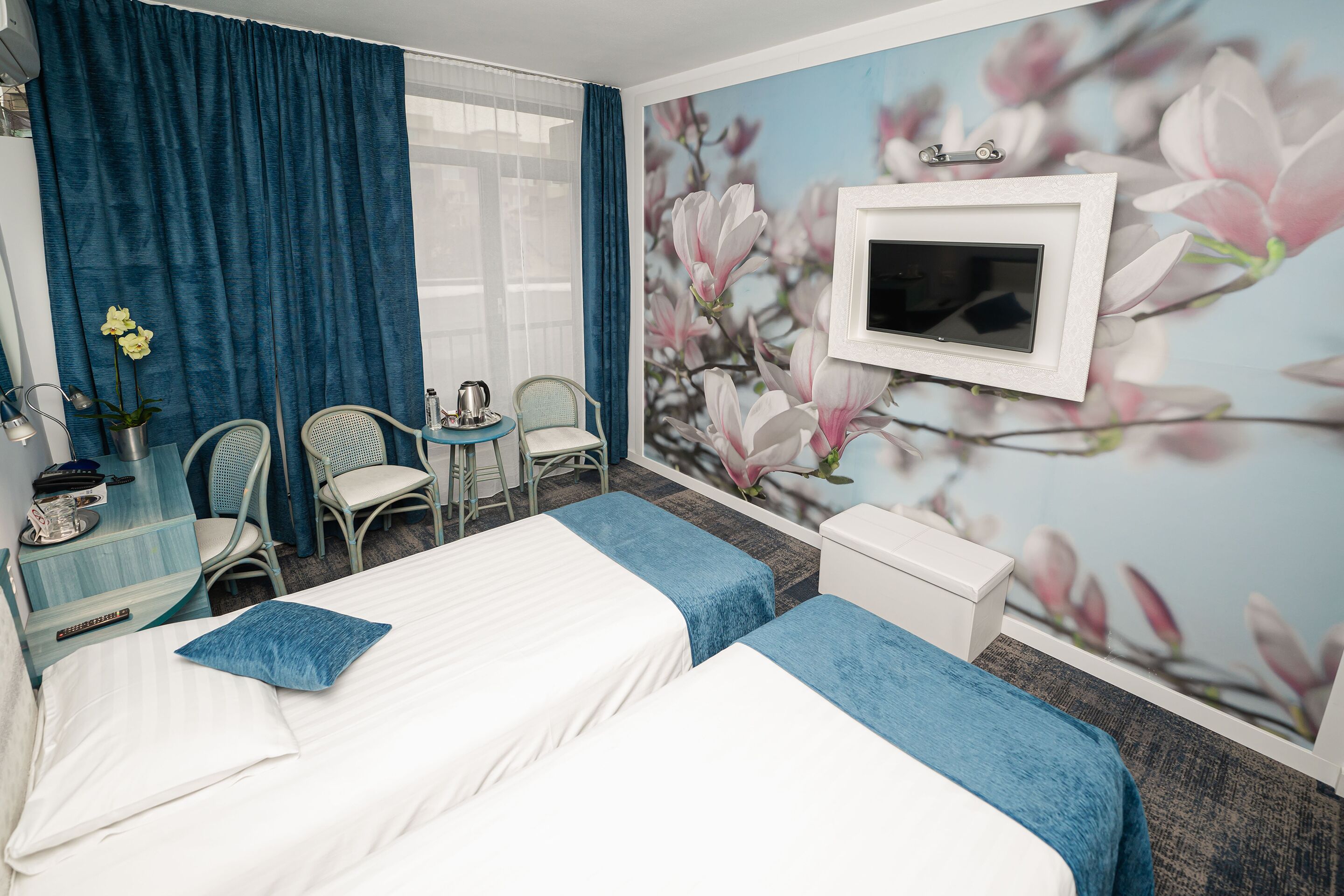 A hotel room with two beds, a TV, a desk with a chair, and a wall with a floral mural.