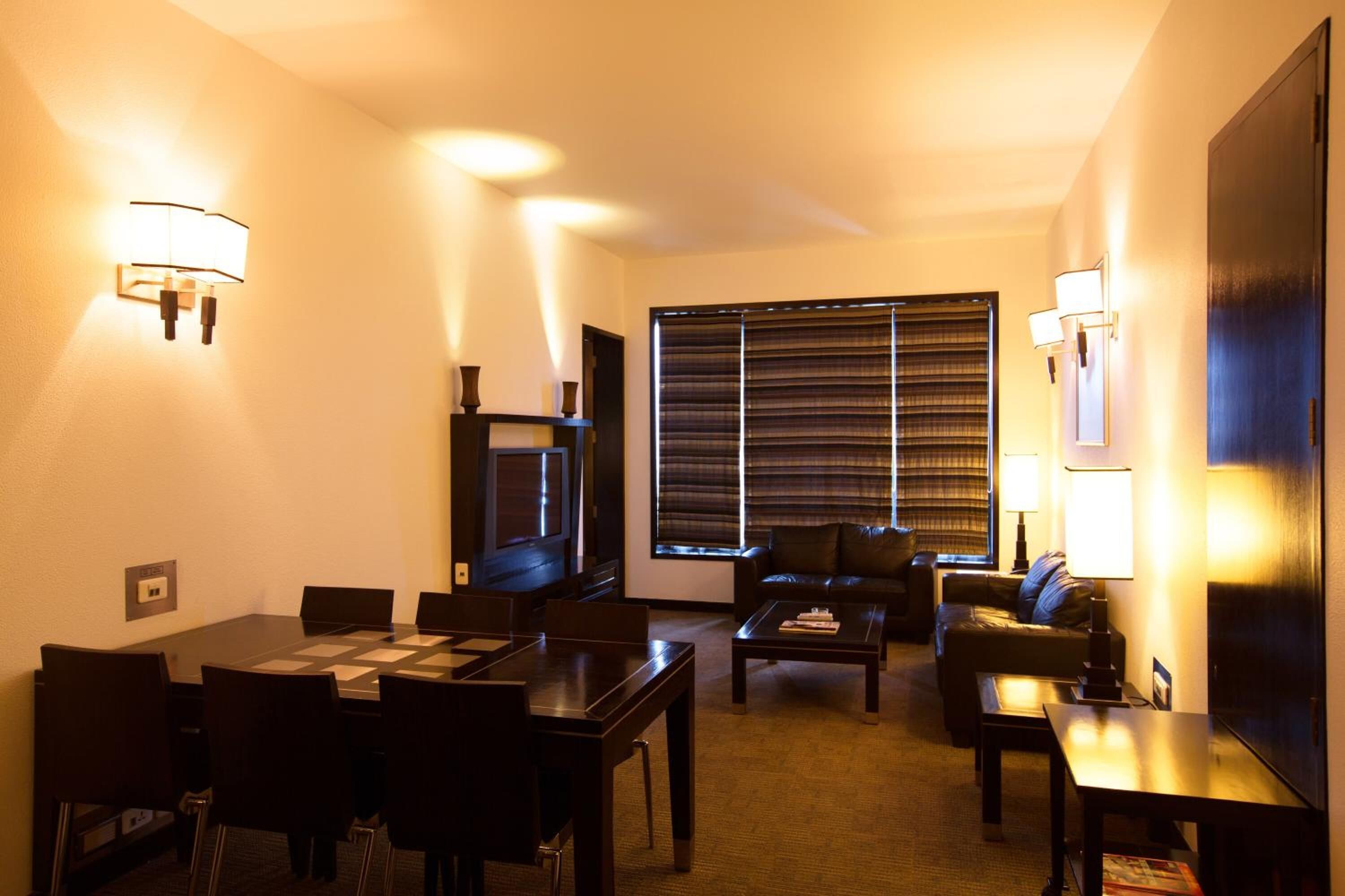 A hotel room with a dining table, chairs, a sofa, a TV, and a window with blinds.