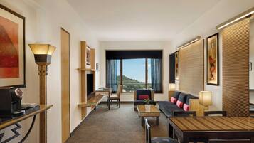 Habitación de hotel con sofá, una mesa de comedor, una cafetera, televisor y un gran ventanal con vistas al paisaje.