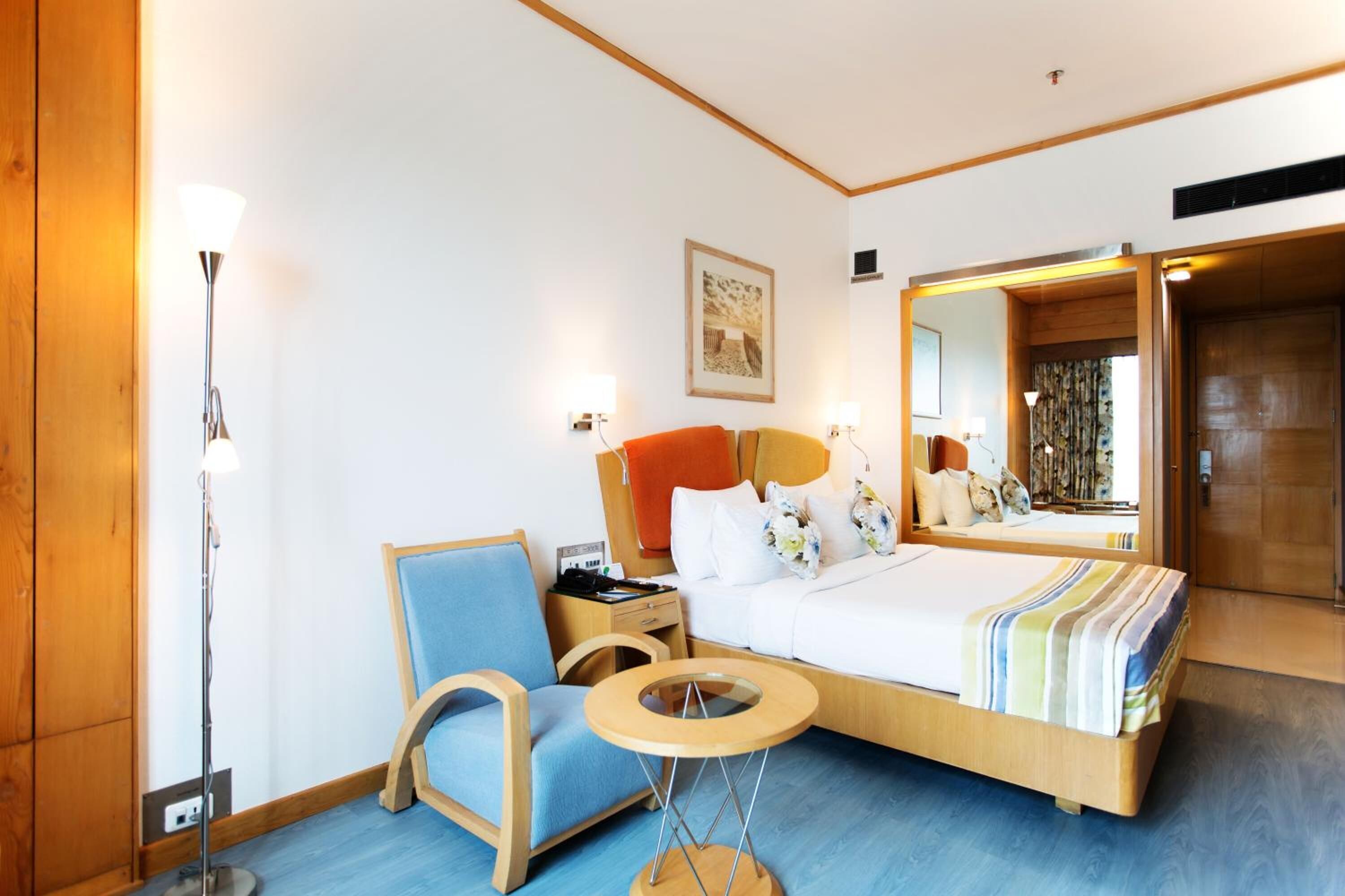 Ett hotellrum med en säng, en stol, ett litet bord, en golvlampa och en spegel.