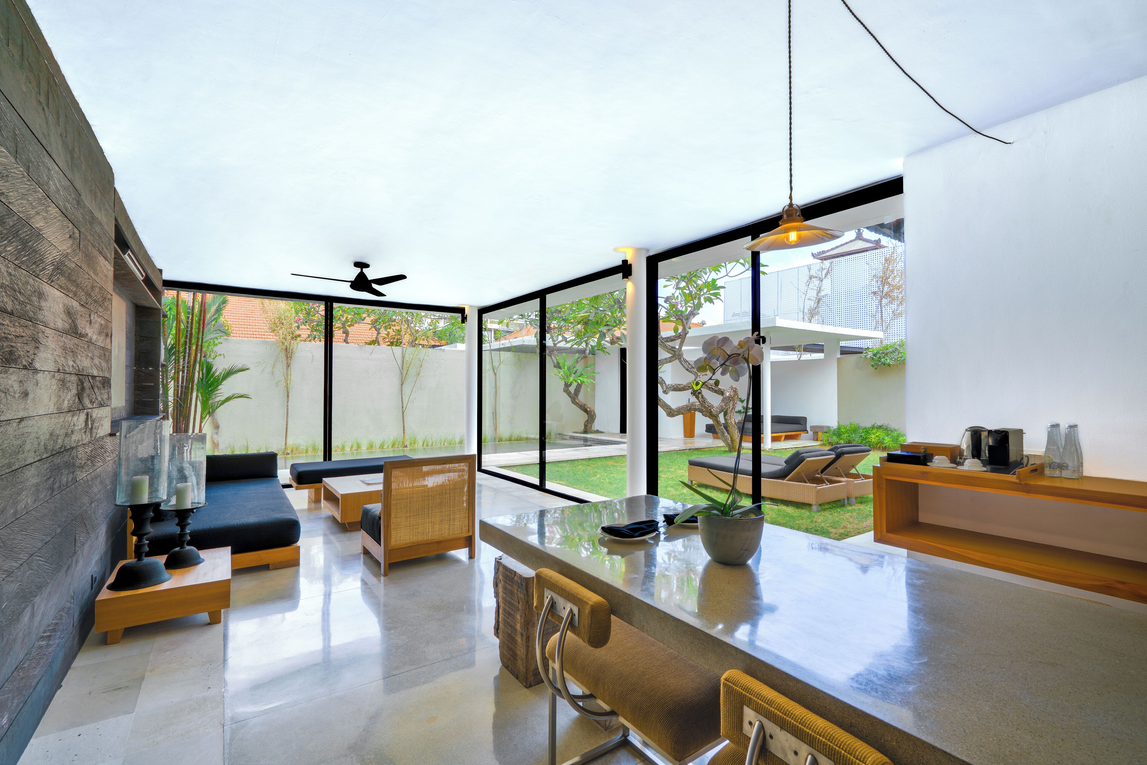 A modern living room with a glass wall, a ceiling fan, and a dining area with a bar counter.